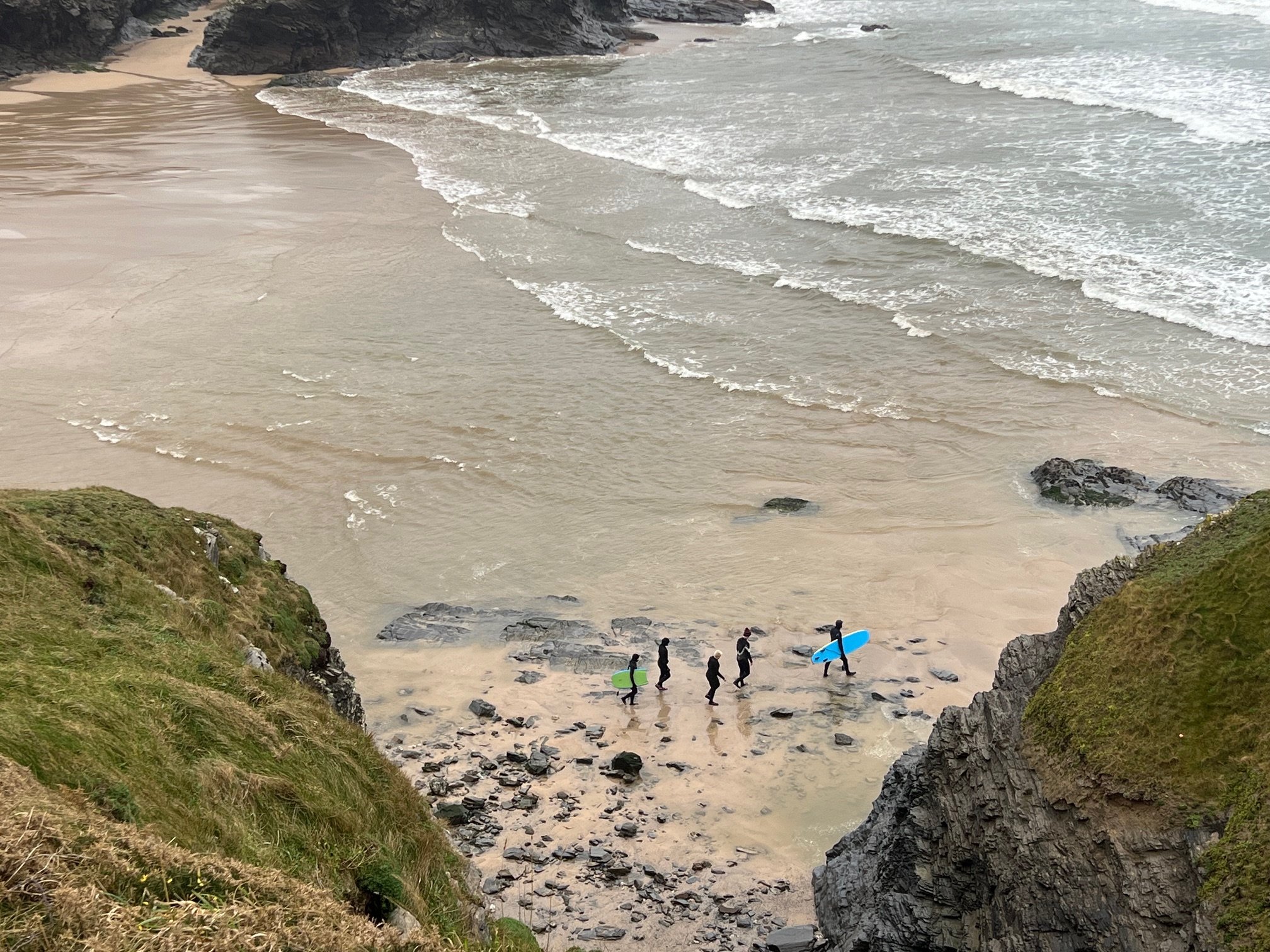 Surfing beach with three surfers carrying boards walking along the shoreline, surrounded by rocky cliffs and waves.