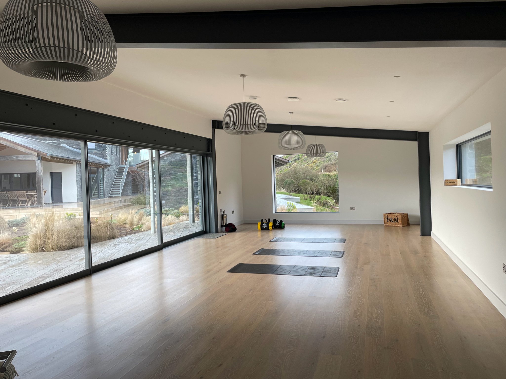 Modern indoor gym with large windows, wooden floor, exercise mats, kettlebells, and contemporary ceiling lights.