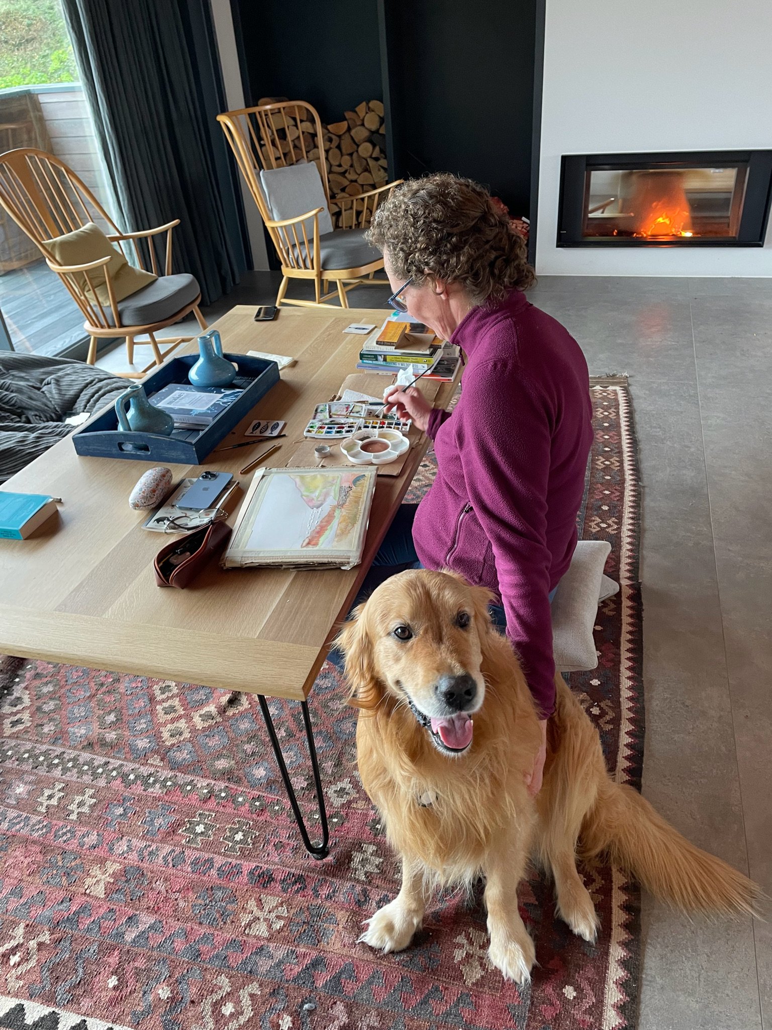 A person in a purple sweater is painting with watercolors at a table, while sitting on a cushion. A golden retriever is sitting beside them, on a patterned rug. The room features wooden chairs, stacked firewood, and a fireplace with a fire burning in