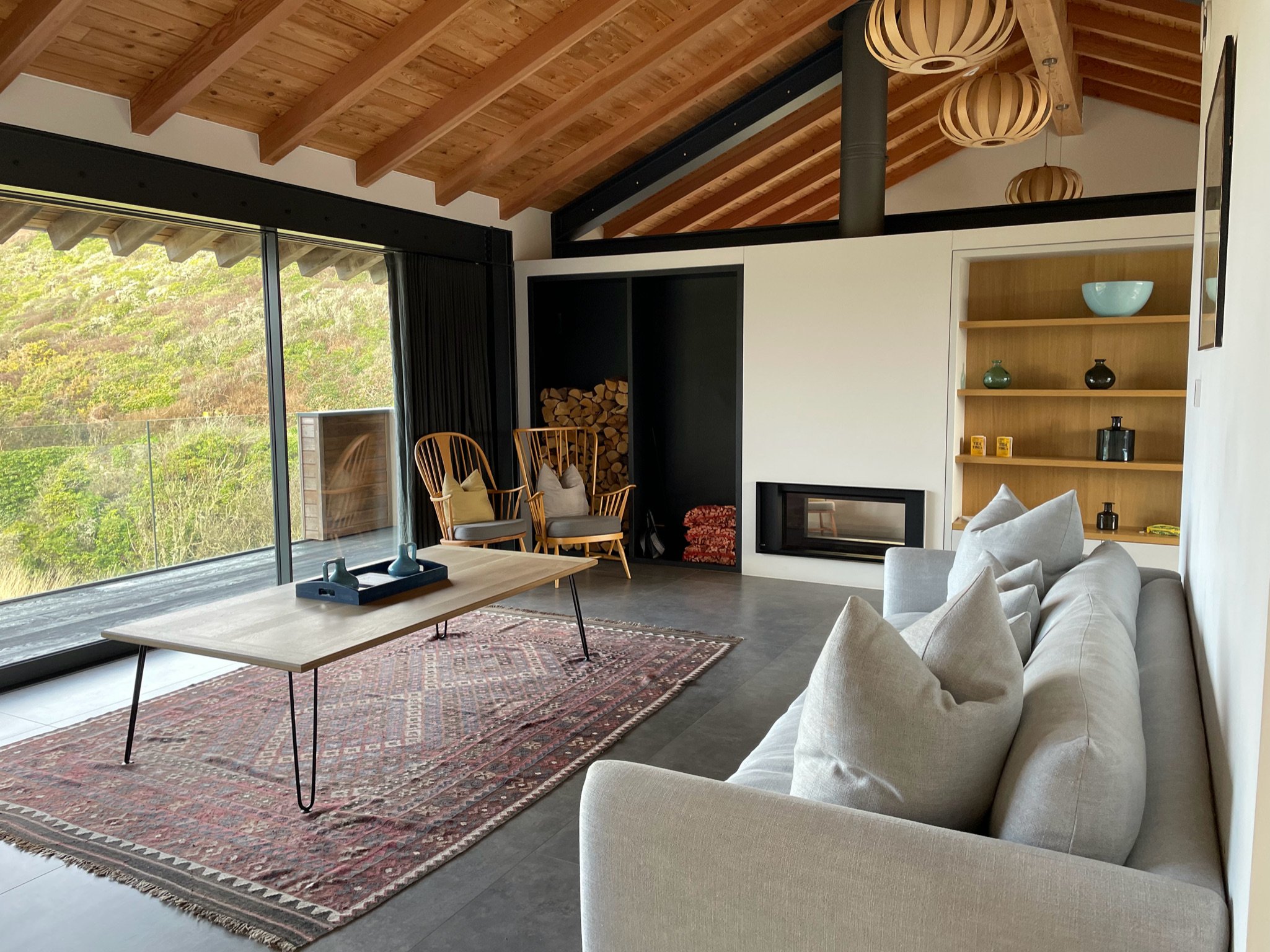 Modern living room with glass walls, gray sofa, coffee table on a patterned rug, wooden chairs, fireplace, and open shelves.