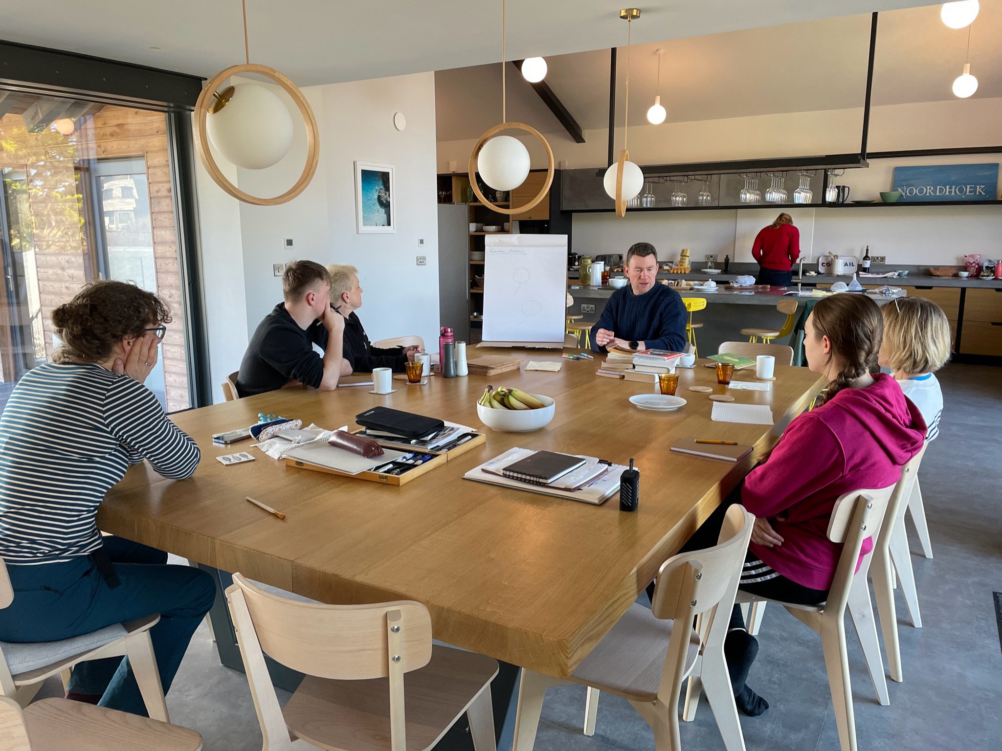 People having a meeting around a wooden table in a modern kitchen setting. The table is filled with notebooks, cups, and plates. A person is cooking in the background.