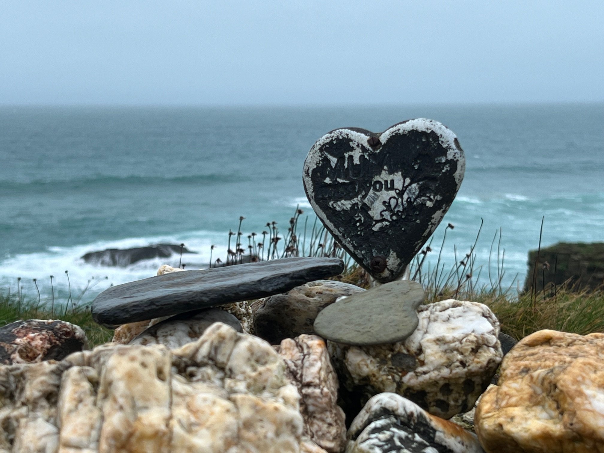 Heart-shaped marker among rocks near a coastline with ocean waves in the background.