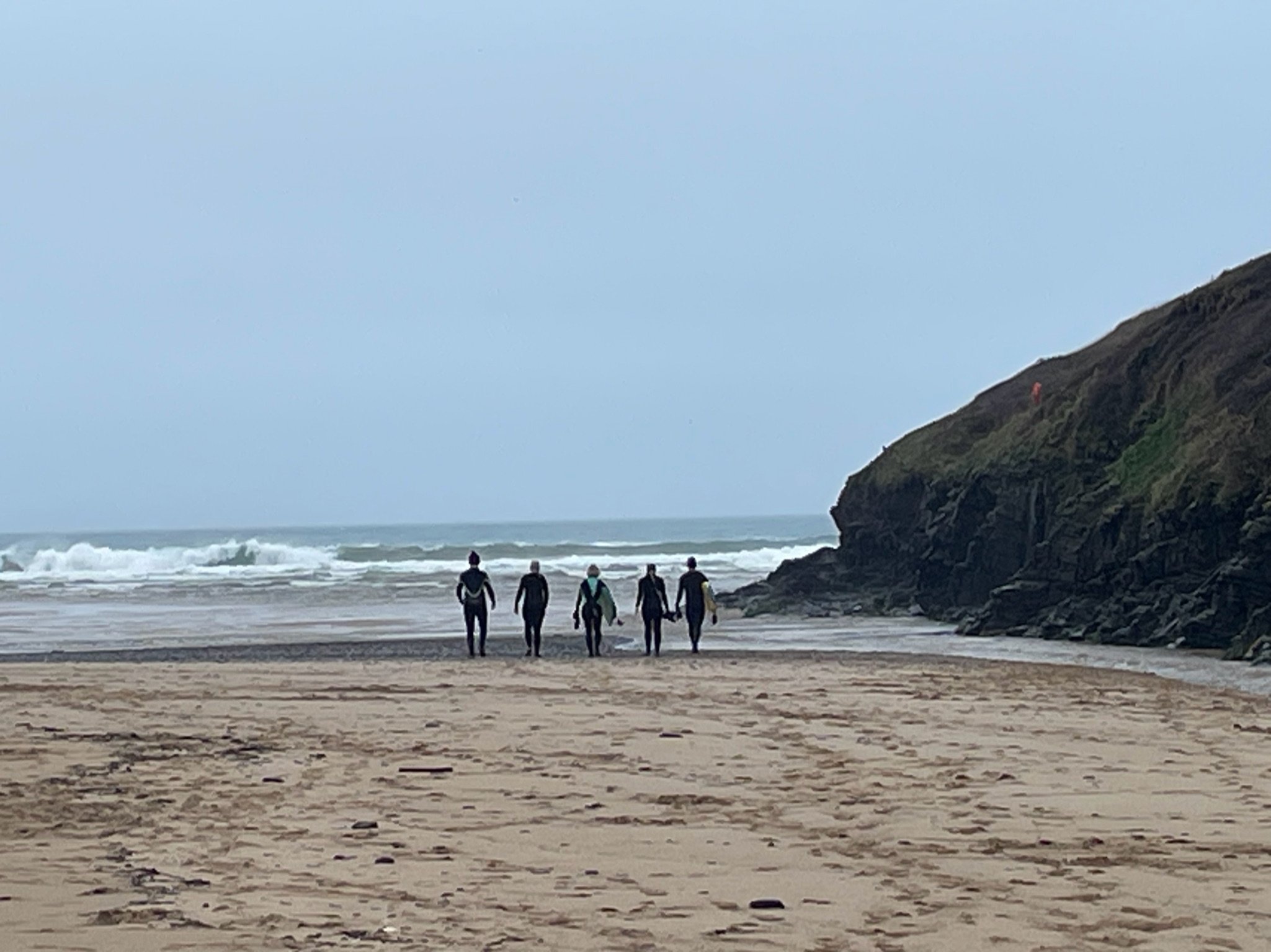 Four people in wetsuits walking on a sandy beach towards the ocean with waves, near rocky cliffs.