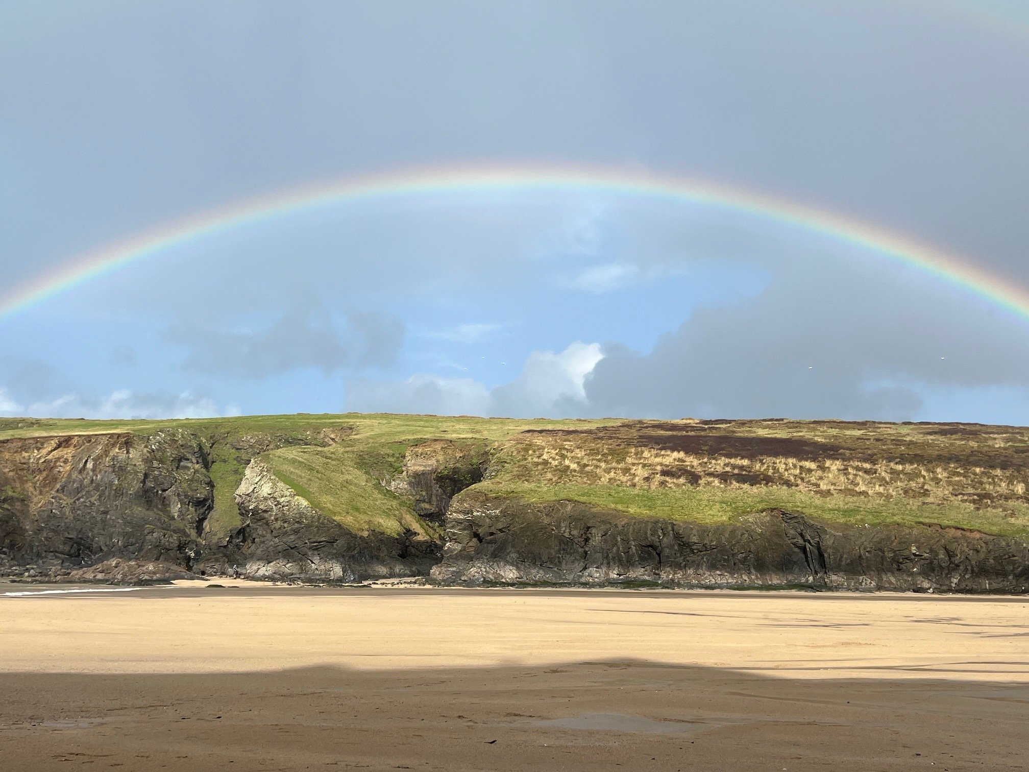 Rainbow over a sandy beach with rocky cliffs and green grass.