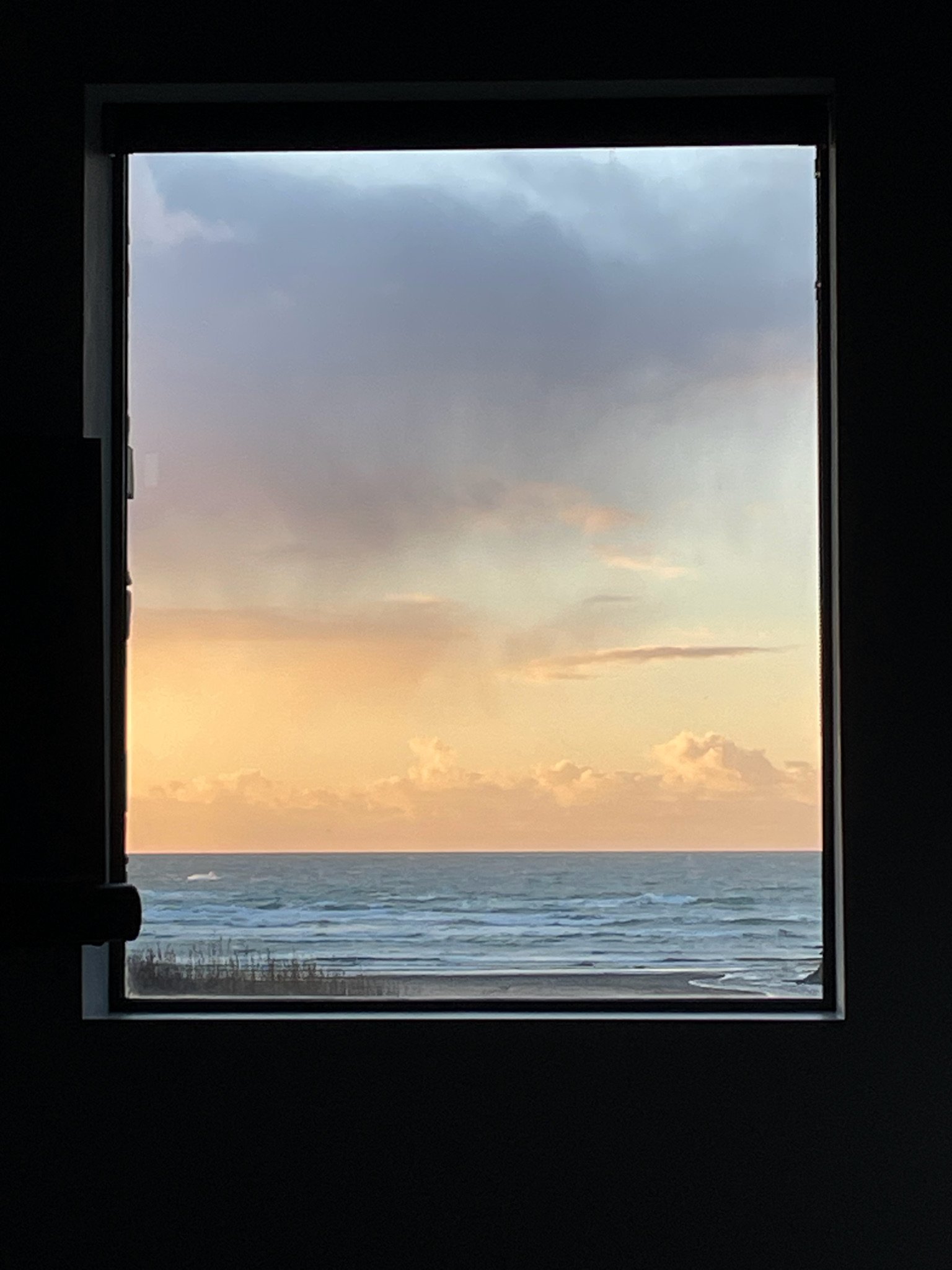 View of ocean and sky through a window, featuring a sunset with clouds and waves on a beach.