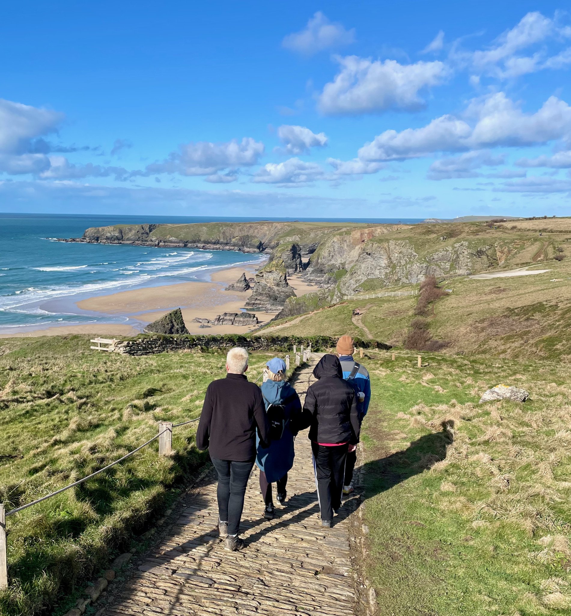 Group of people walking on a coastal path overlooking cliffs and ocean, with blue skies and scattered clouds in the background.
