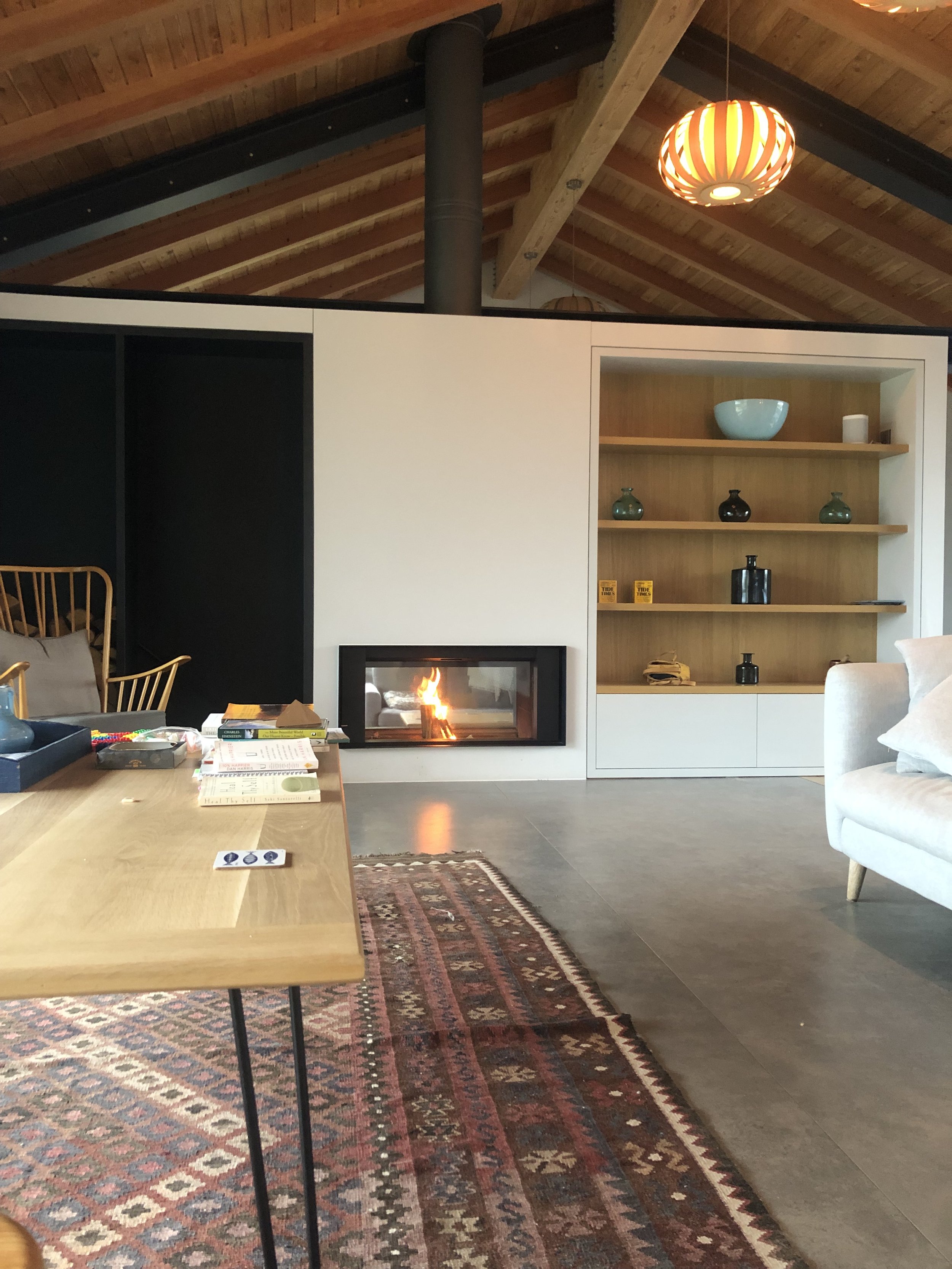 Cozy living room with a modern fireplace, wooden ceiling, and decorative shelves. A wooden table with books and a patterned rug are in the foreground, and there is a gray sofa to the right.