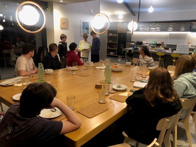 Group of people sitting around a large dining table in a modern kitchen, listening to a presentation or discussion from a few standing individuals. Plates, glasses, and light fixtures are visible.