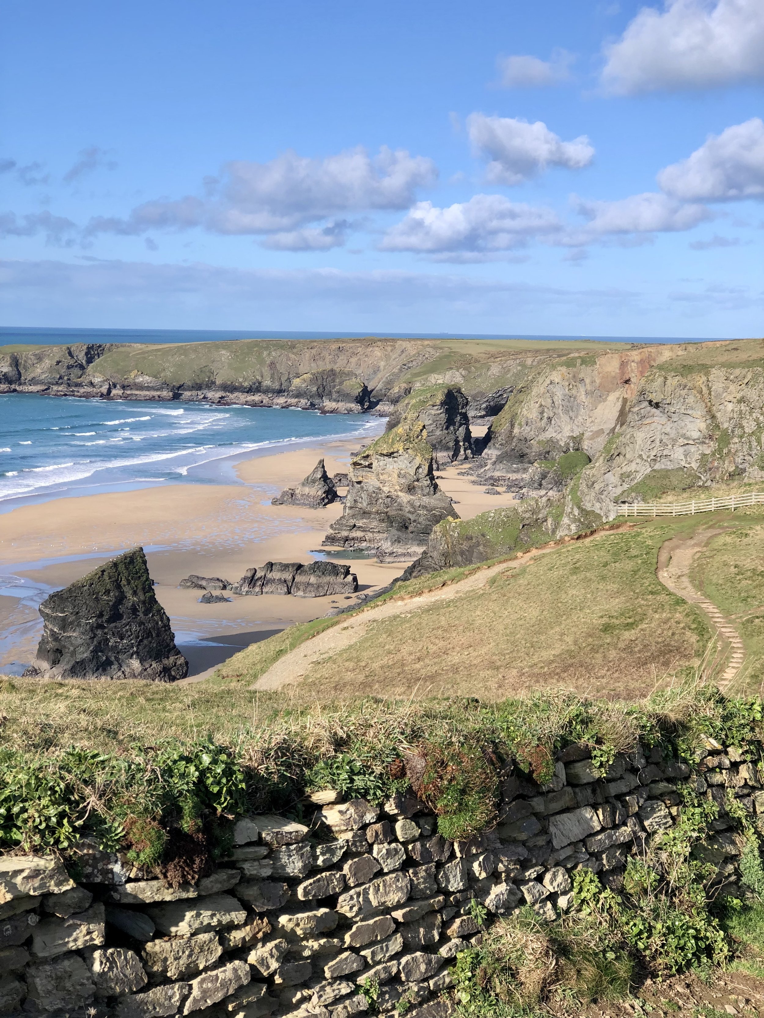 Coastal landscape with rocky cliffs, sandy beach, ocean waves, and stone wall, under a blue sky with fluffy clouds.