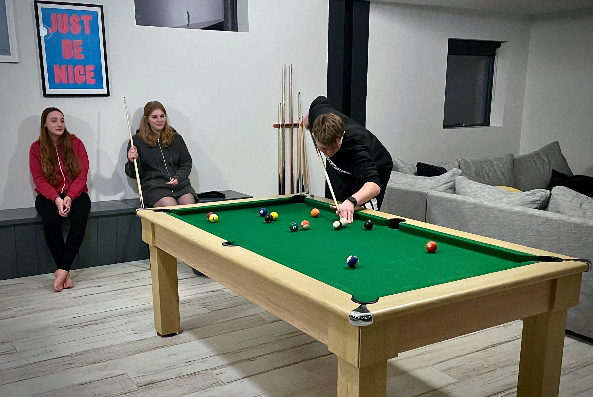 Three people in a room with a pool table. One person is playing pool, while two others are sitting nearby. A "Just Be Nice" poster is on the wall.