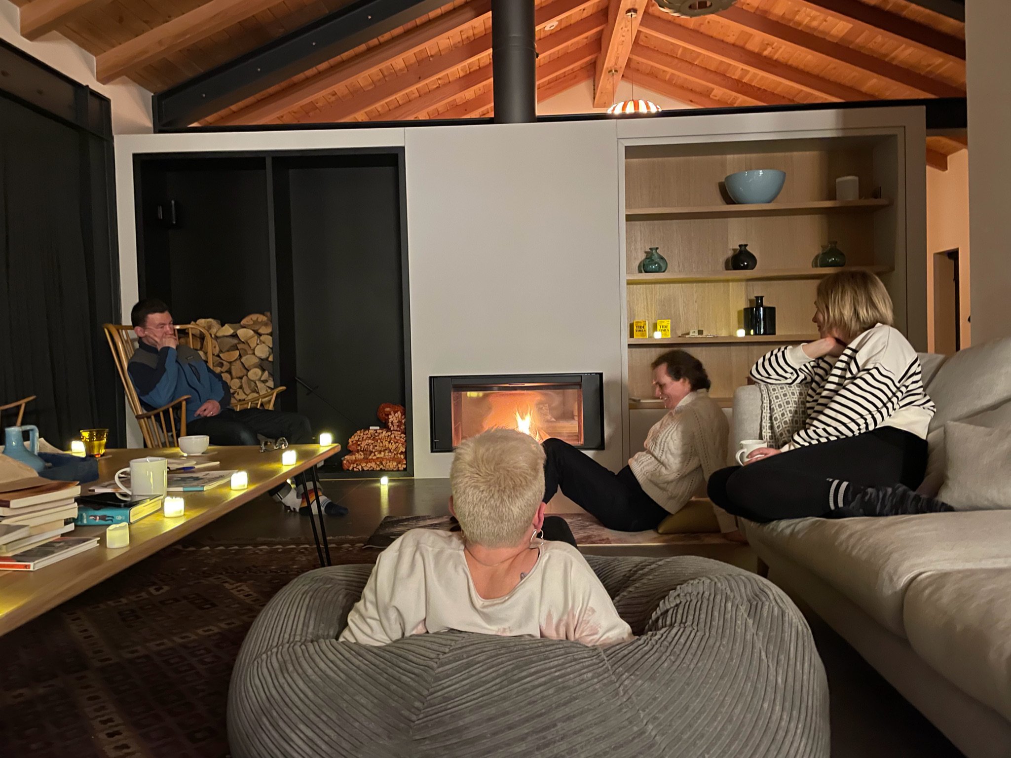 People in a cozy living room, sitting by a modern fireplace. The room has a wooden ceiling and shelves with decorative items. A person on the left is on a chair near stacked firewood, while others sit on a couch and a bean bag. Candles and books on a