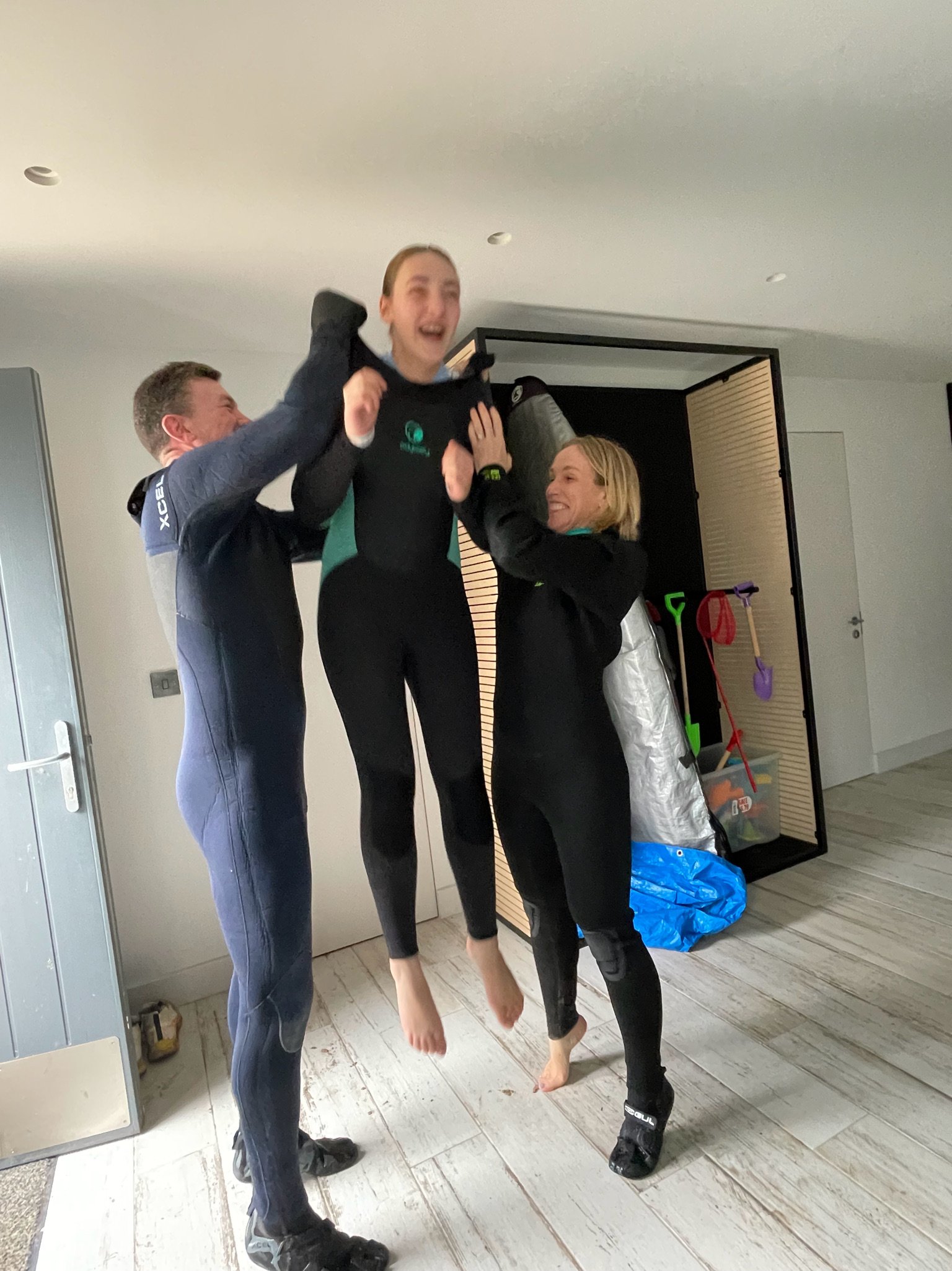 Three people indoors in wetsuits are playfully lifting each other, smiling and having fun.