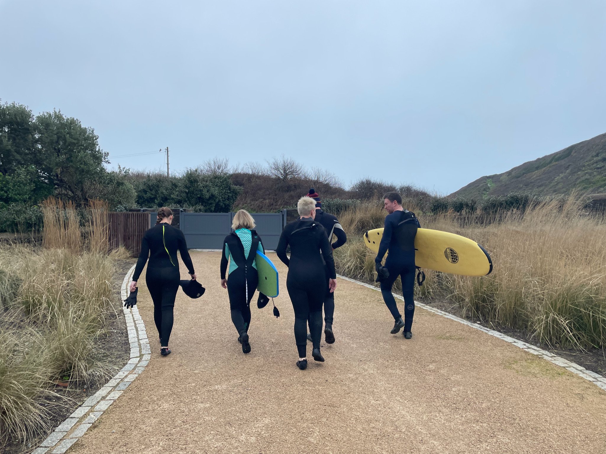 Group of four people in wetsuits walking on a path with surfboards, surrounded by grasses.