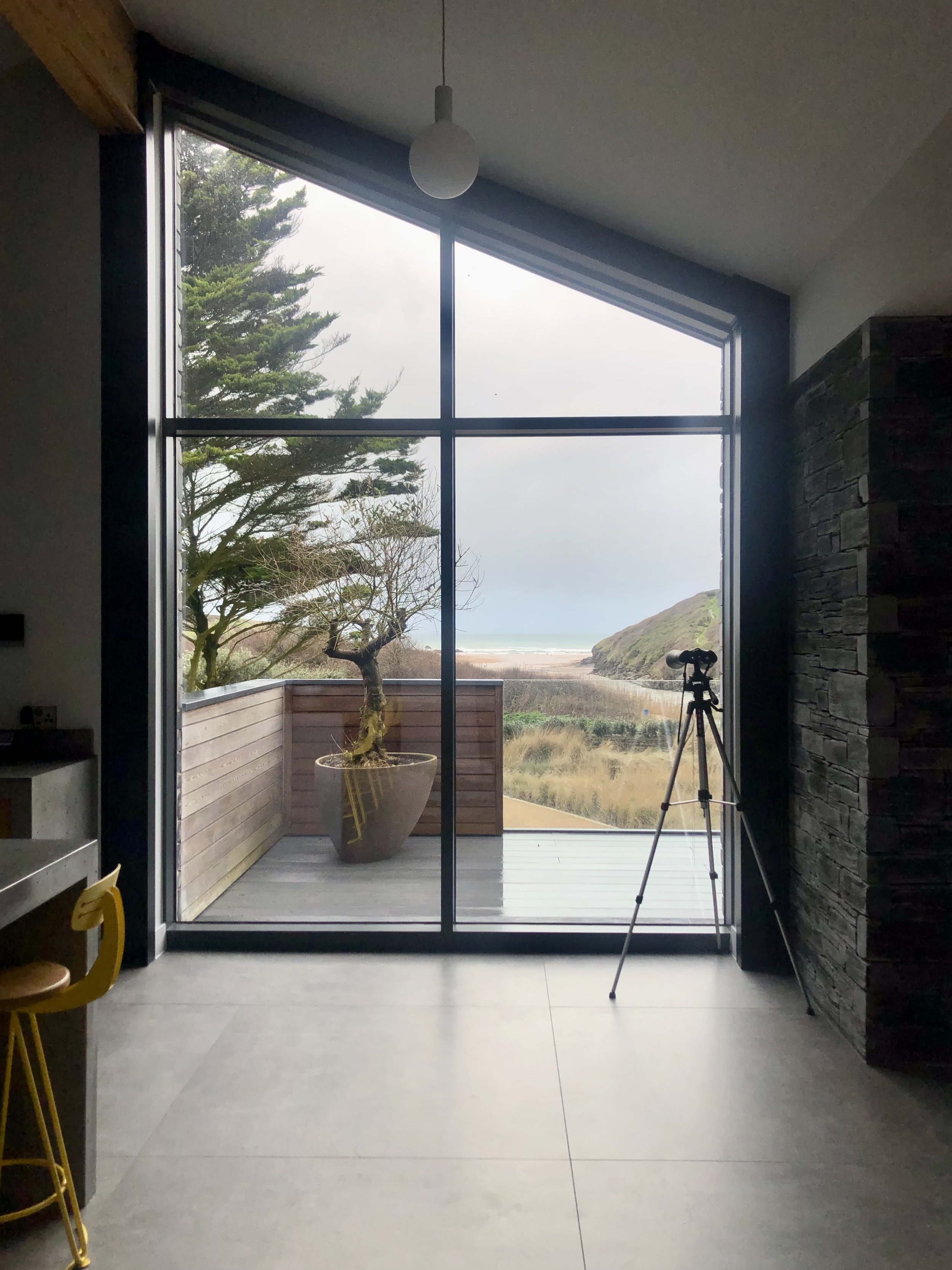 Interior of a modern room with large floor-to-ceiling windows offering a view of a wooden deck, potted tree, tripod, and distant landscape, including hills and a body of water.
