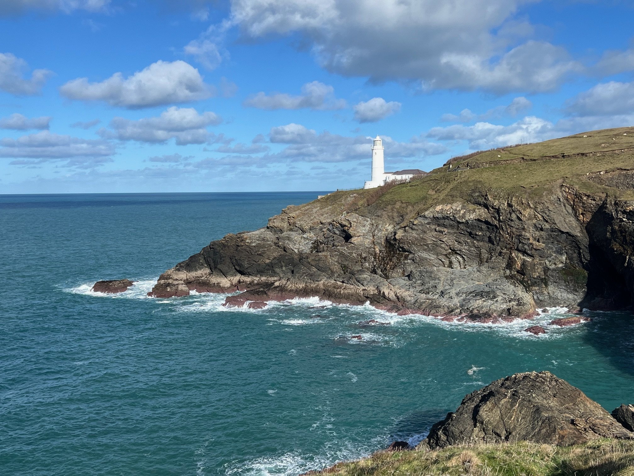 Coastal landscape with a white lighthouse on a rocky cliff, surrounded by the ocean, under a blue sky with scattered clouds.