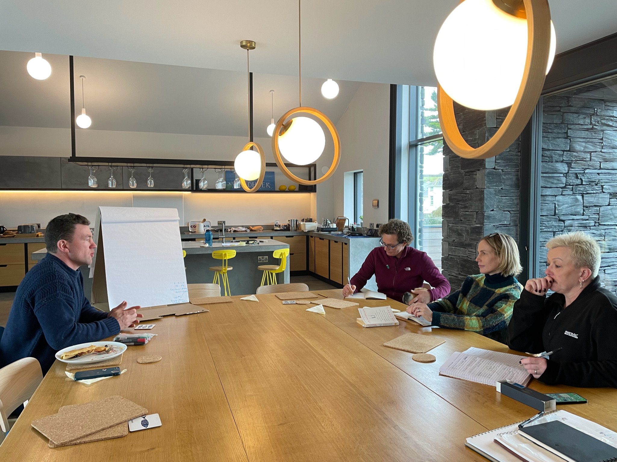 A group of four people sitting around a wooden conference table in a modern, well-lit room with large windows. One person is speaking while others take notes. There is a flip chart, table mats, and various personal items on the table. The room featur
