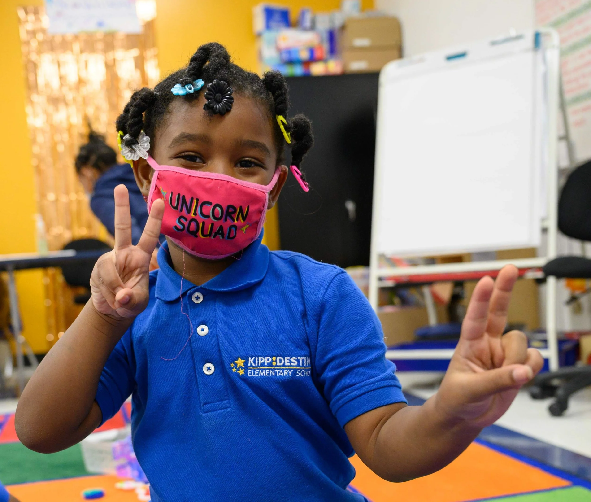 A Black girl in a blue polo shirt makes peace signs while wearing a mask that reads "Unicorn Squad"