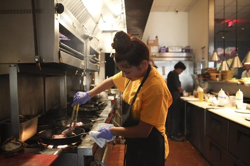 A girl prepares food in an industrial kitchen while wearing a black apron