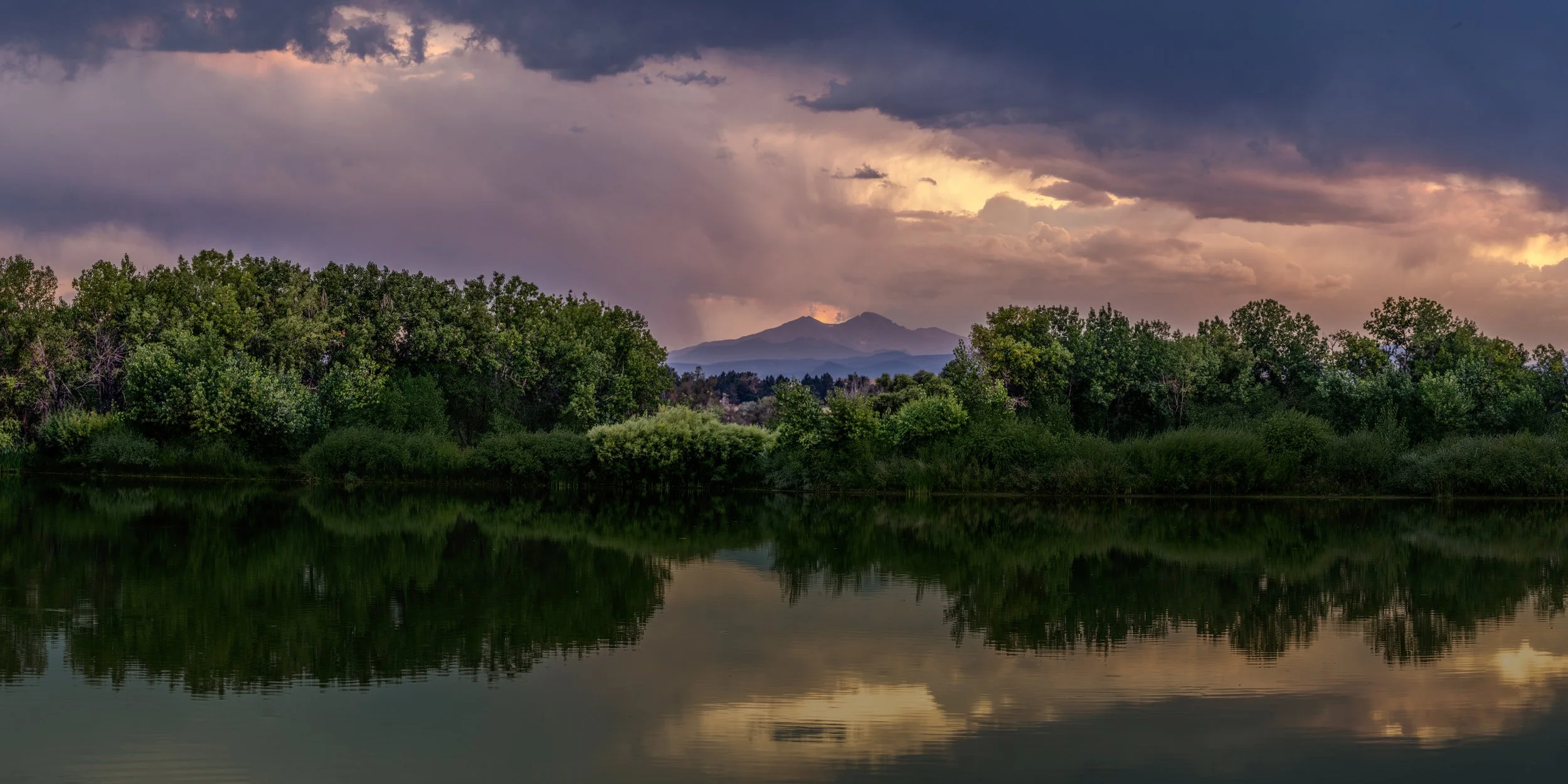 Photo of storm clouds gathering over Arapaho Bends pond with Longs Peak and Mount Meeker in the background.