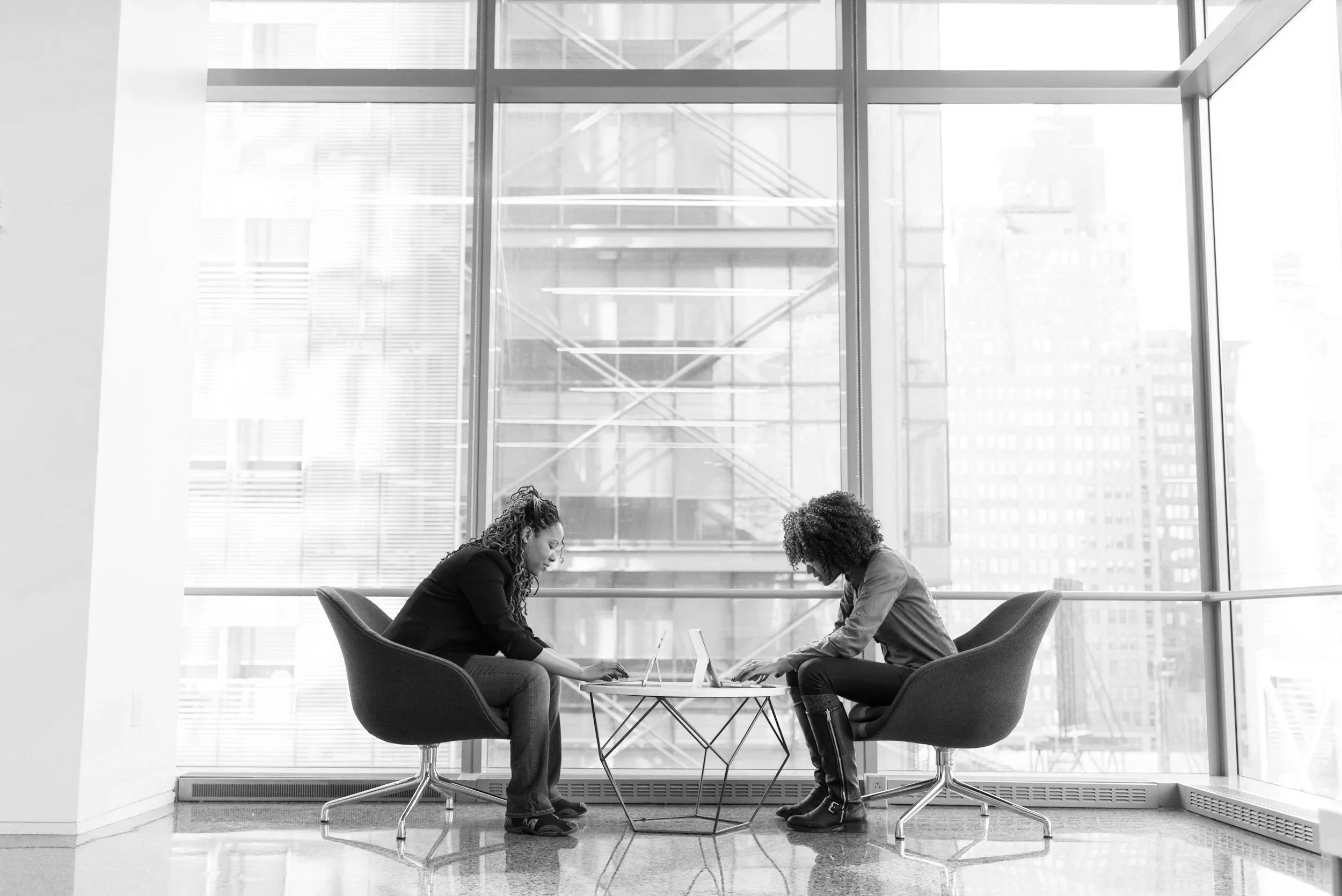 Two women sitting at a round glass table in front of large windows overlooking city buildings, engaged in conversation or work.