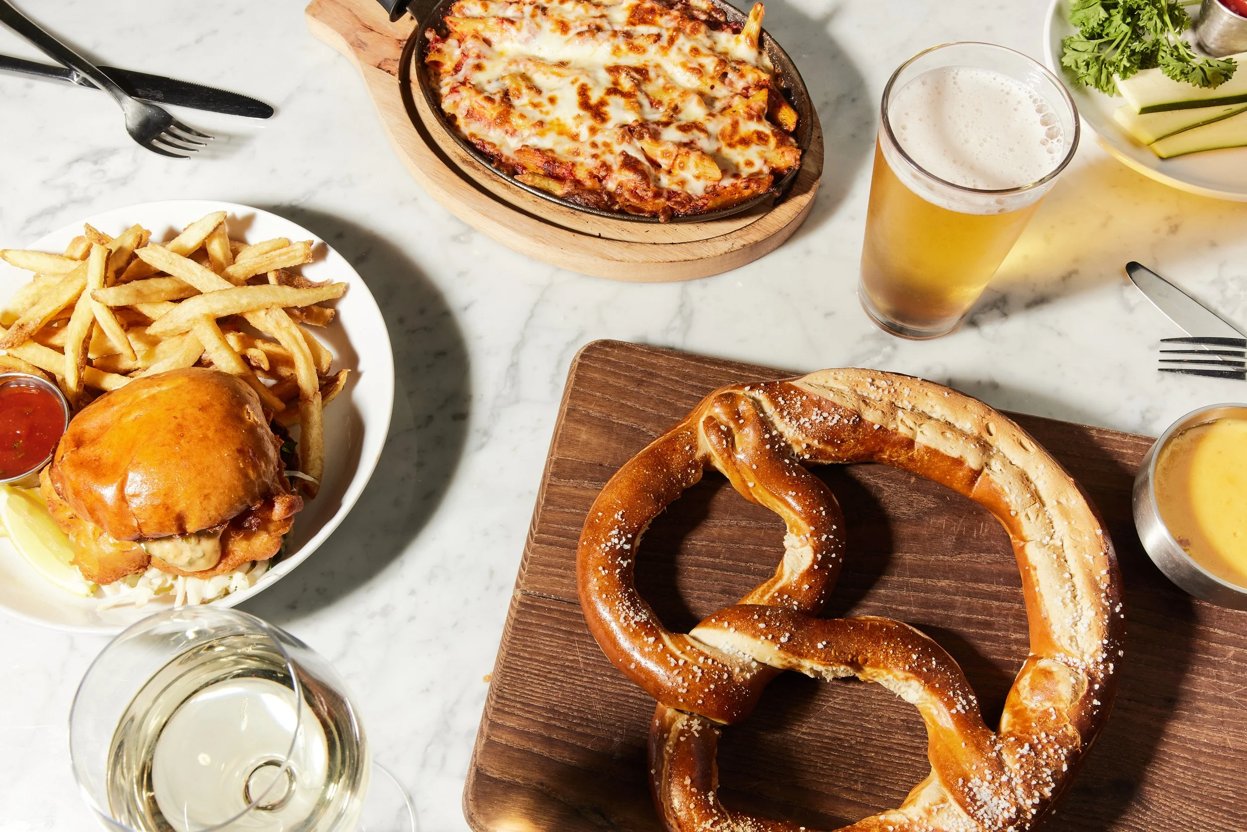 A spread of food on a white marble table, including a plate of French fries with a small cup of ketchup, a cheeseburger with lettuce on a white plate, a large pretzel on a wooden board, a glass of white wine, a glass of beer, a bowl of cheese sauce, 