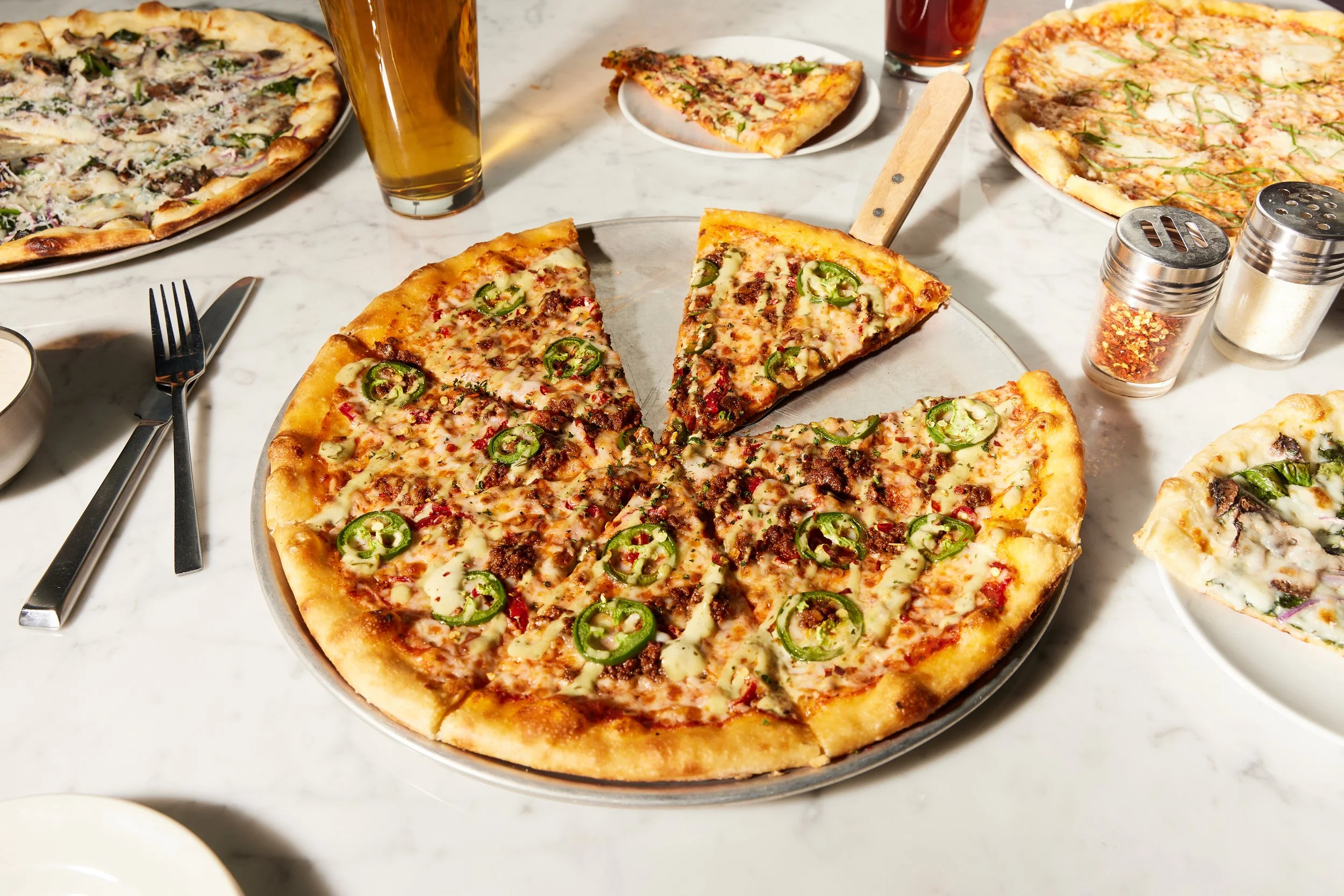 A table with three pizzas, two glasses of beer, a small bowl of sauce, and salt and pepper shakers. One pizza is topped with jalapeños and ground beef, and the others have various toppings.