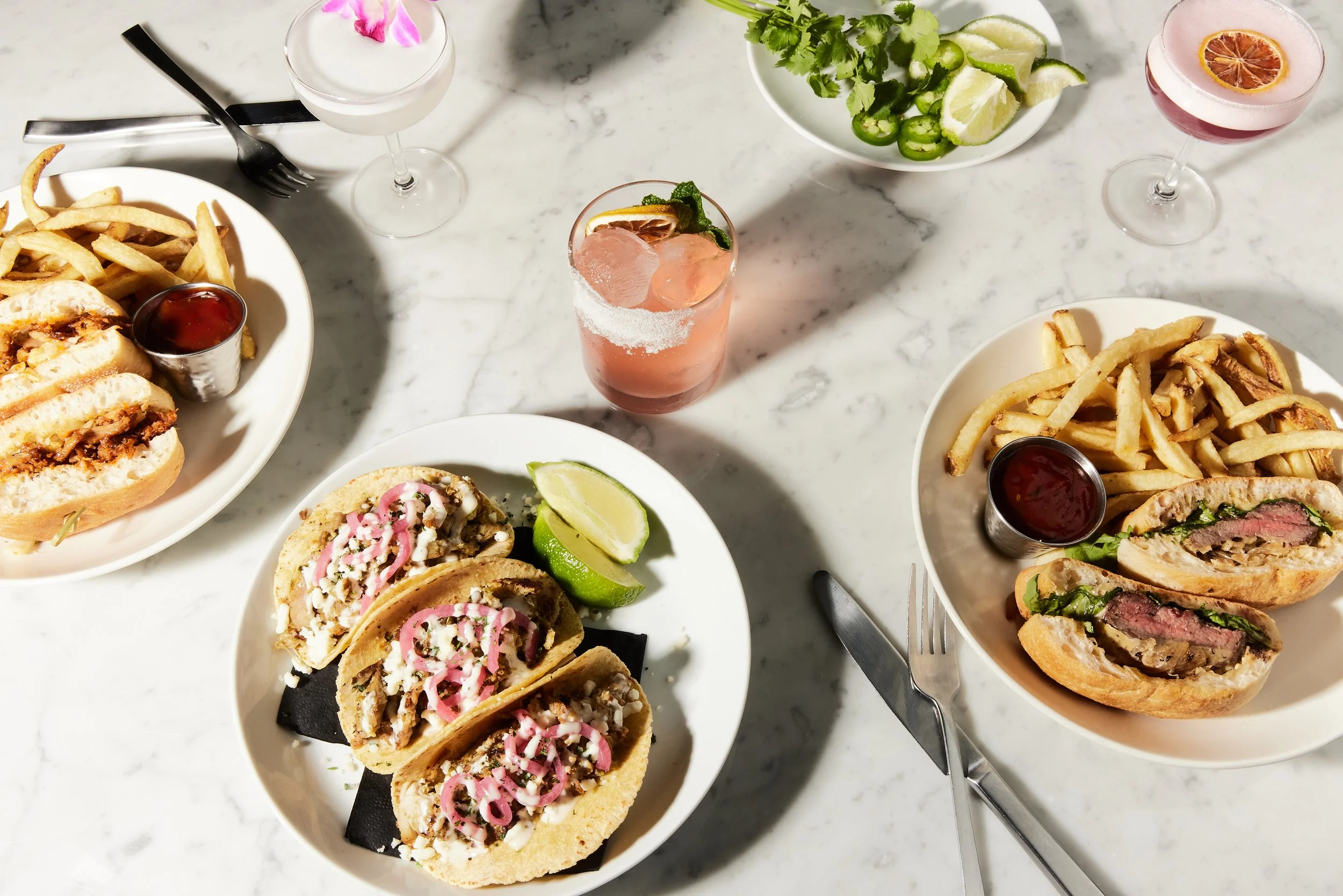 Overhead view of two plates with open-faced sandwiches and fries, three cocktails, and a bowl of lime wedges and jalapeños on a white marble table.