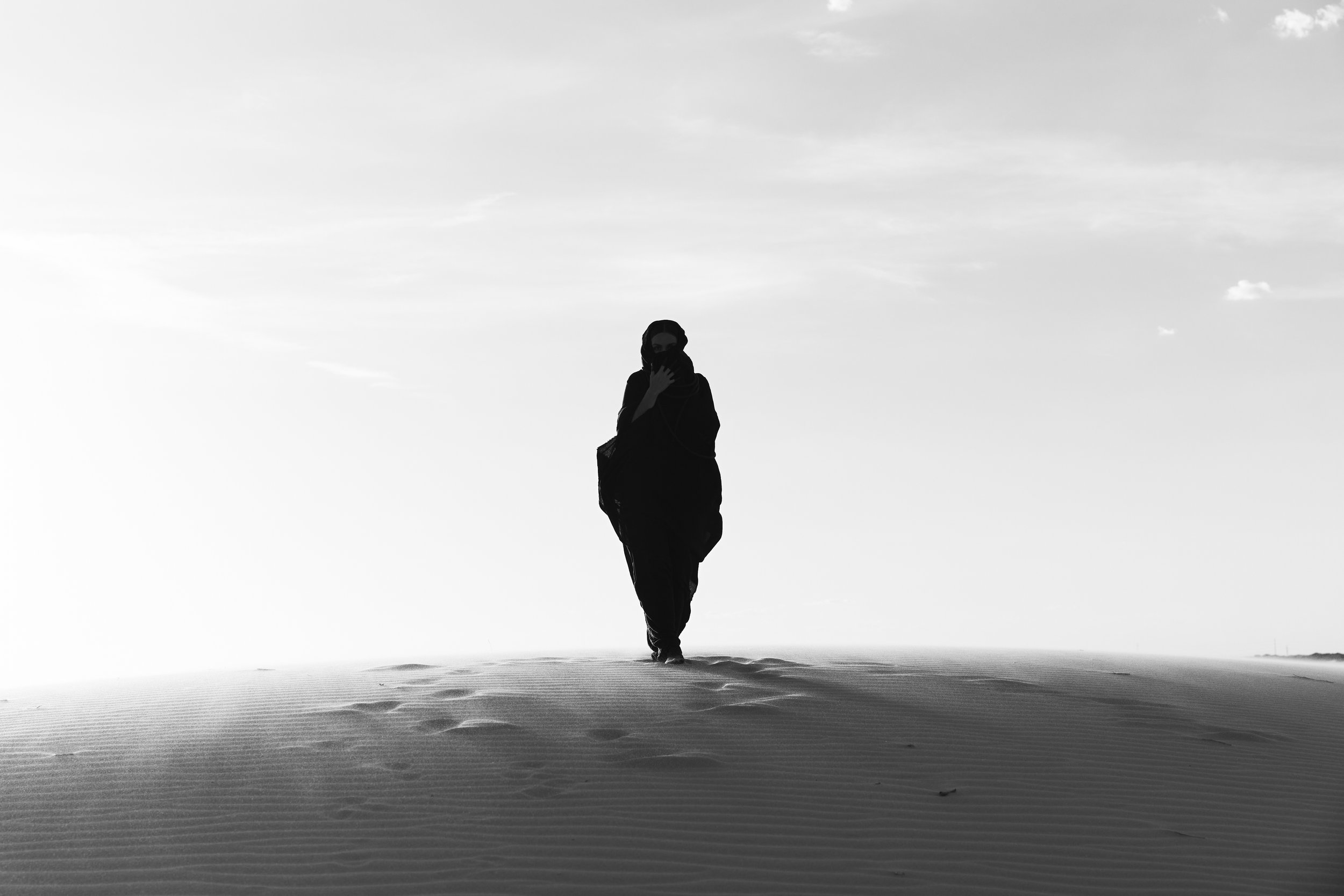 A person in dark clothing walking through a desert, arm raised to their face, with a cloudy sky overhead.