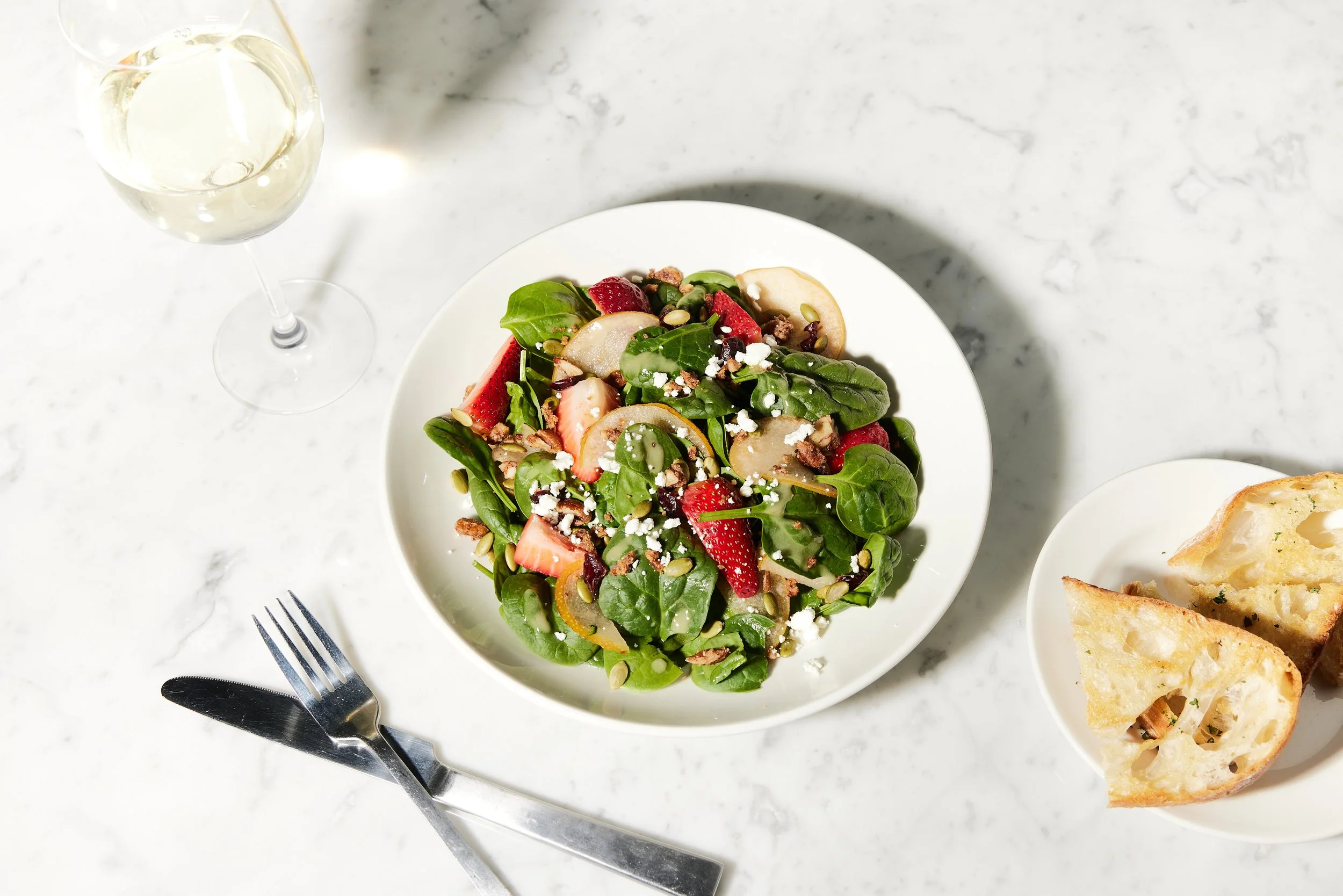 A plate of mixed salad with strawberries, yellow peppers, greens, nuts, and cheese, next to halves of toasted bread on a small plate, with a glass of white wine and a fork and knife on a white marble surface.