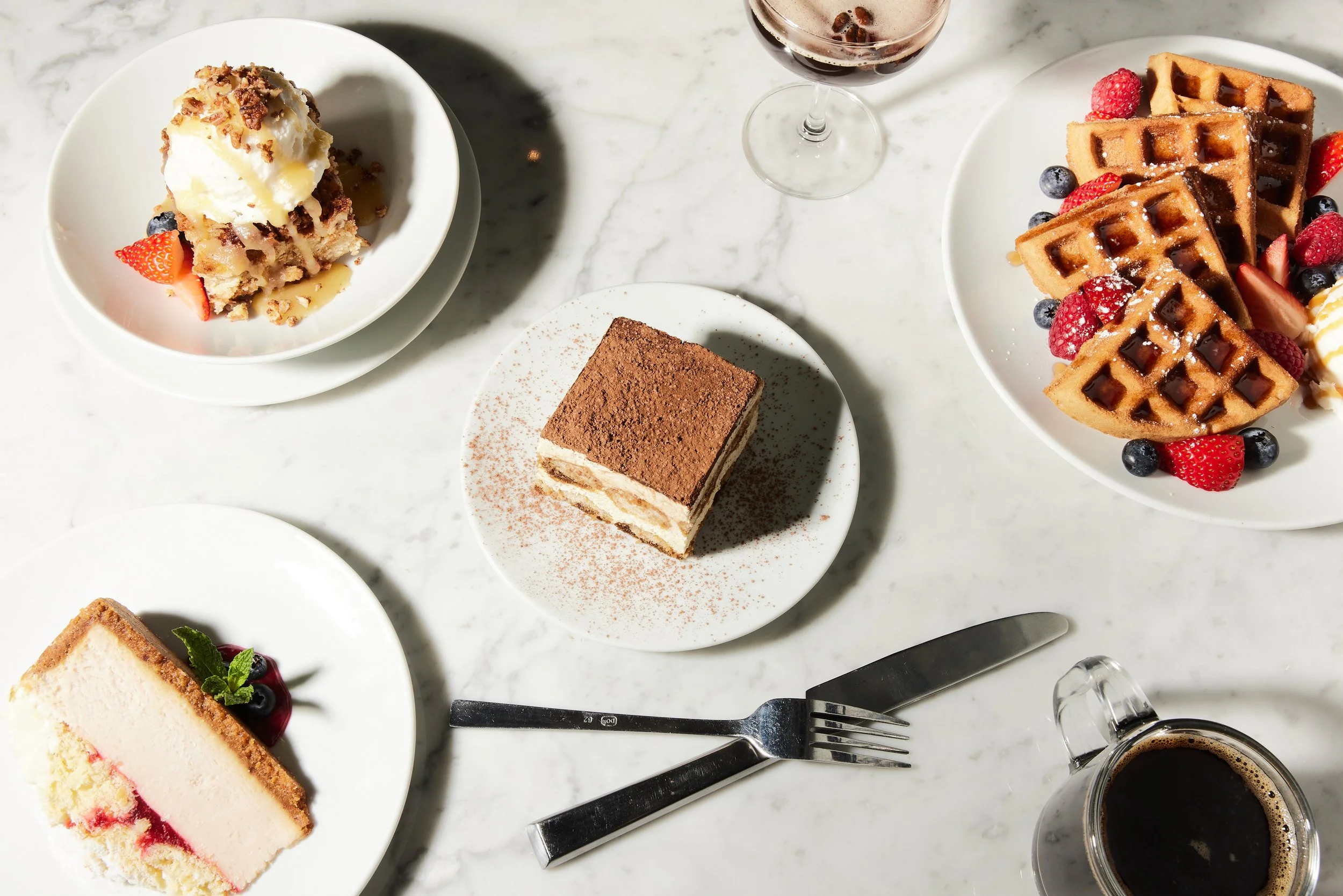 Assorted desserts including a slice of cheesecake with berries, a tiramisu, waffles with berries, a scoop of ice cream, and a glass of coffee on a white marble table.