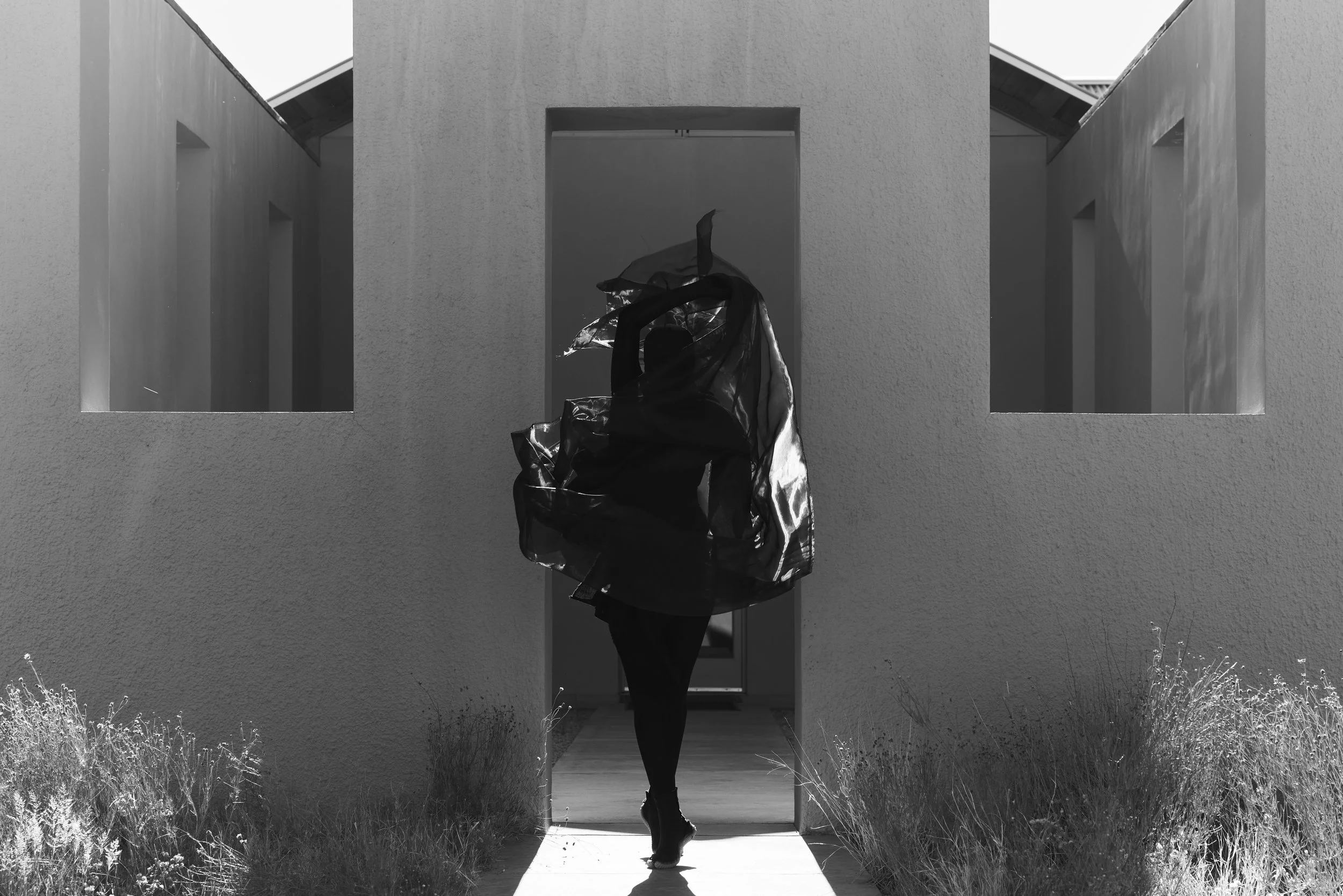 A person walking through a small opening in a modern building with textured walls, wearing a dark outfit and carrying a shiny, transparent bag.