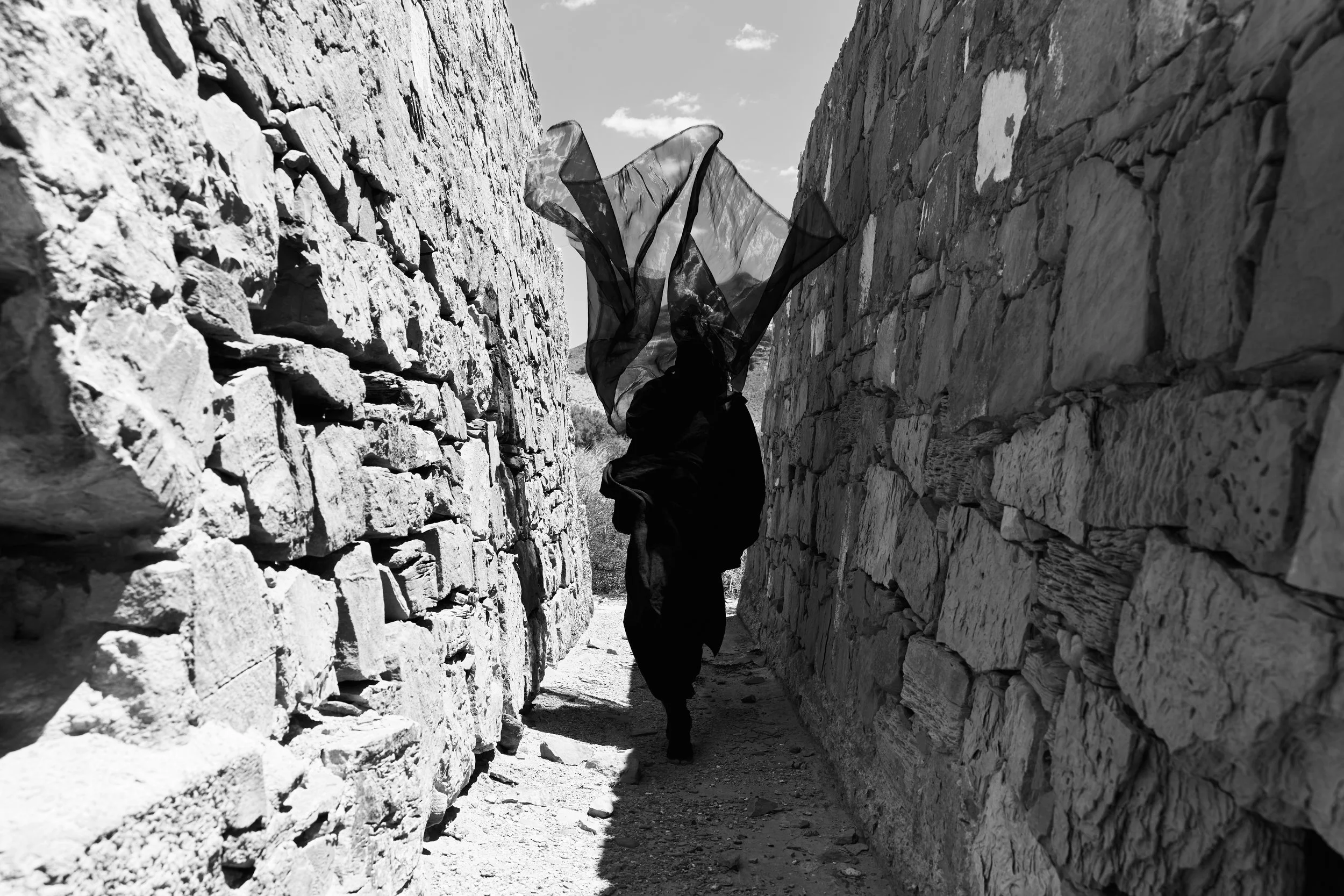 A person walking through a narrow stone passageway, holding a flowing black scarf in the air. The passage is flanked by tall stone walls, and the scene is in black and white.
