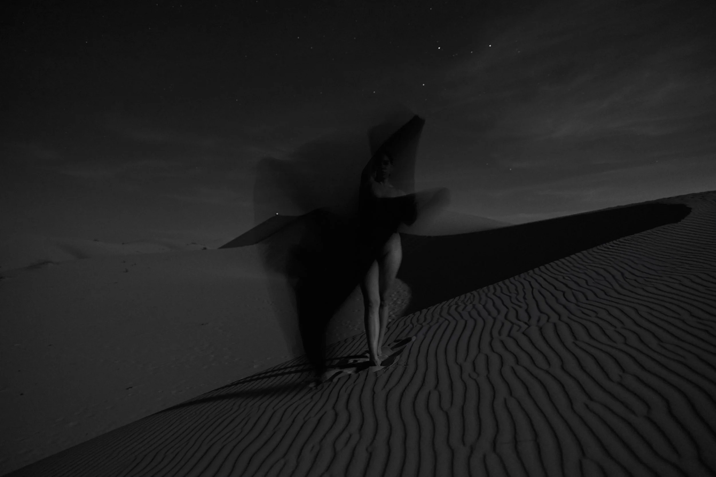 A woman standing in a desert at night, holding a large mysterious object creating a shadow on the sand.