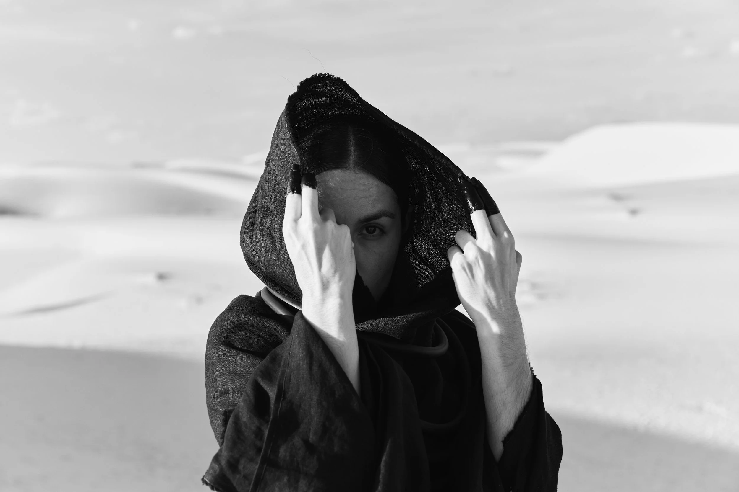 A person with dark hair wearing black clothing and a hood, holding the hood with both hands, standing in a sandy desert landscape under a cloudy sky.