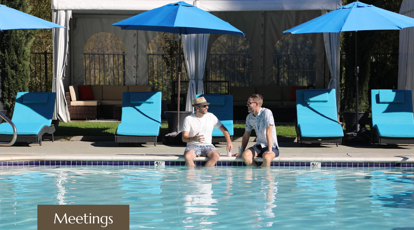 Two men sitting at the edge of a swimming pool, engaged in conversation, with pool chairs and umbrellas in the background.