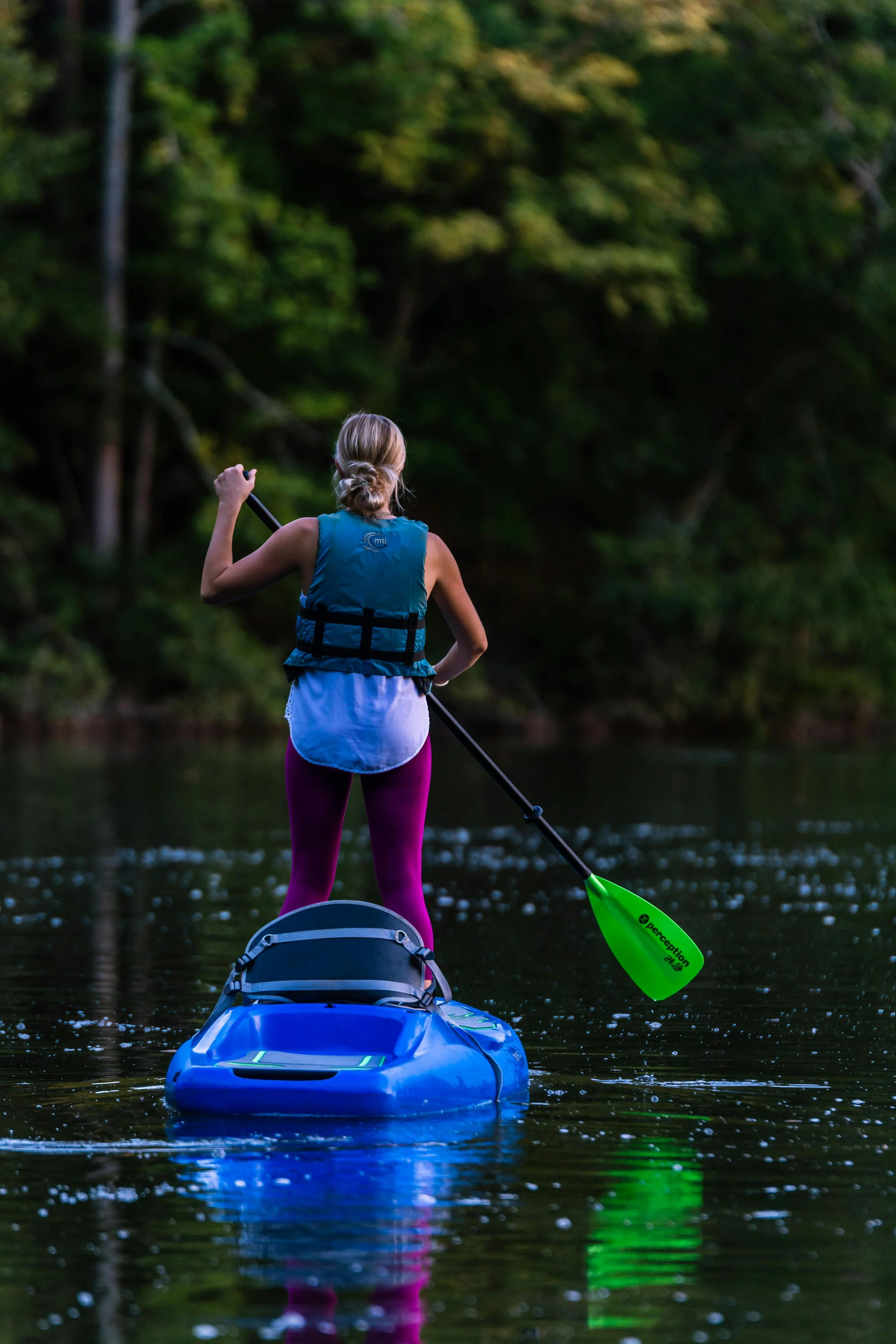 Woman paddleboarding on a calm river surrounded by lush green trees, wearing a blue life jacket and pink leggings.