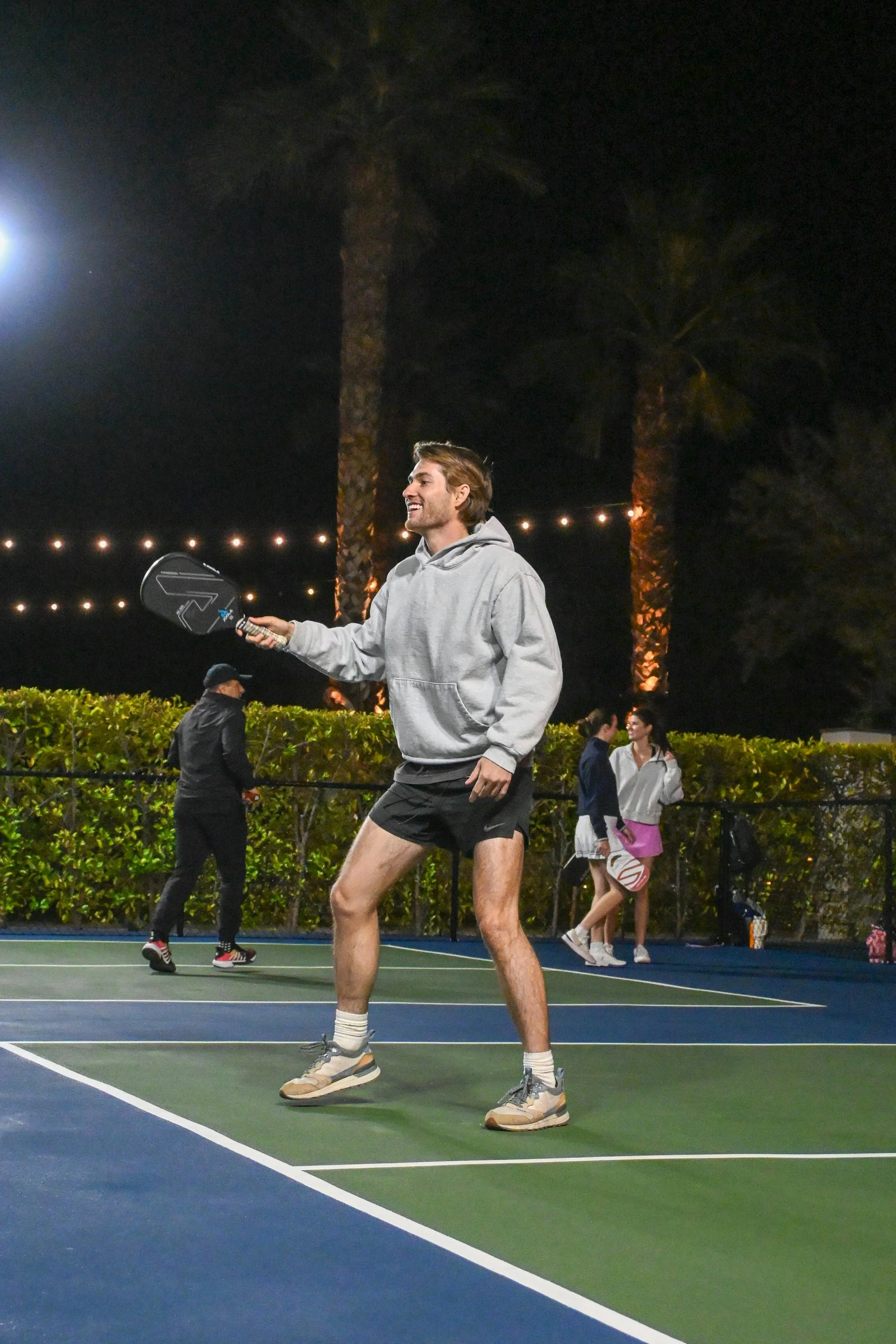 Young man playing pickleball at night on an outdoor court surrounded by palm trees and string lights, with other players in the background.
