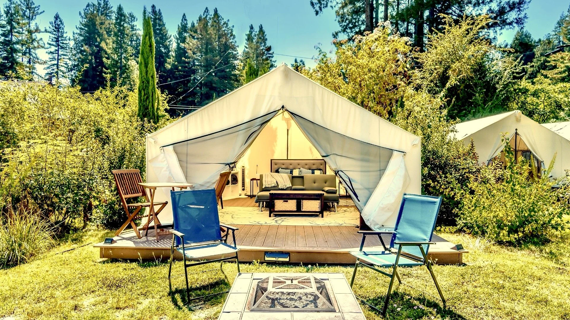 A glamping tent set up on a wooden platform in a lush, green wooded area with seating and interior furnishings visible