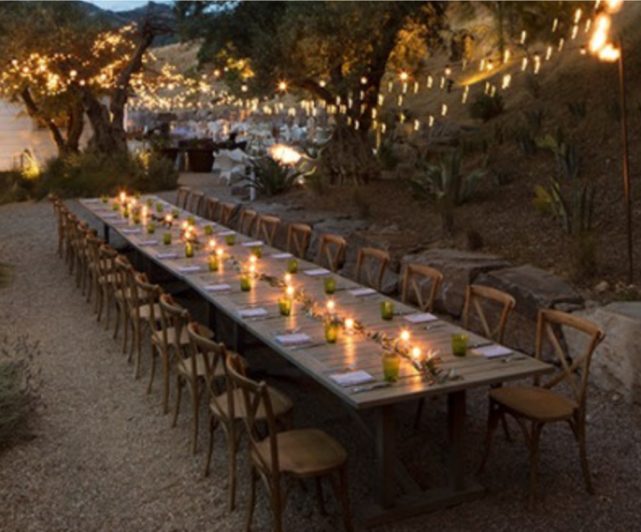 Long outdoor dining table set for a dinner celebration with candles and small potted plants, under string lights in a garden at dusk.