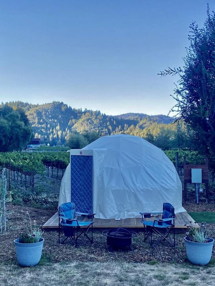 A yurt set on a wooden platform in a garden with two blue camping chairs, potted plants, and a fire pit, surrounded by greenery and hills under a clear blue sky.