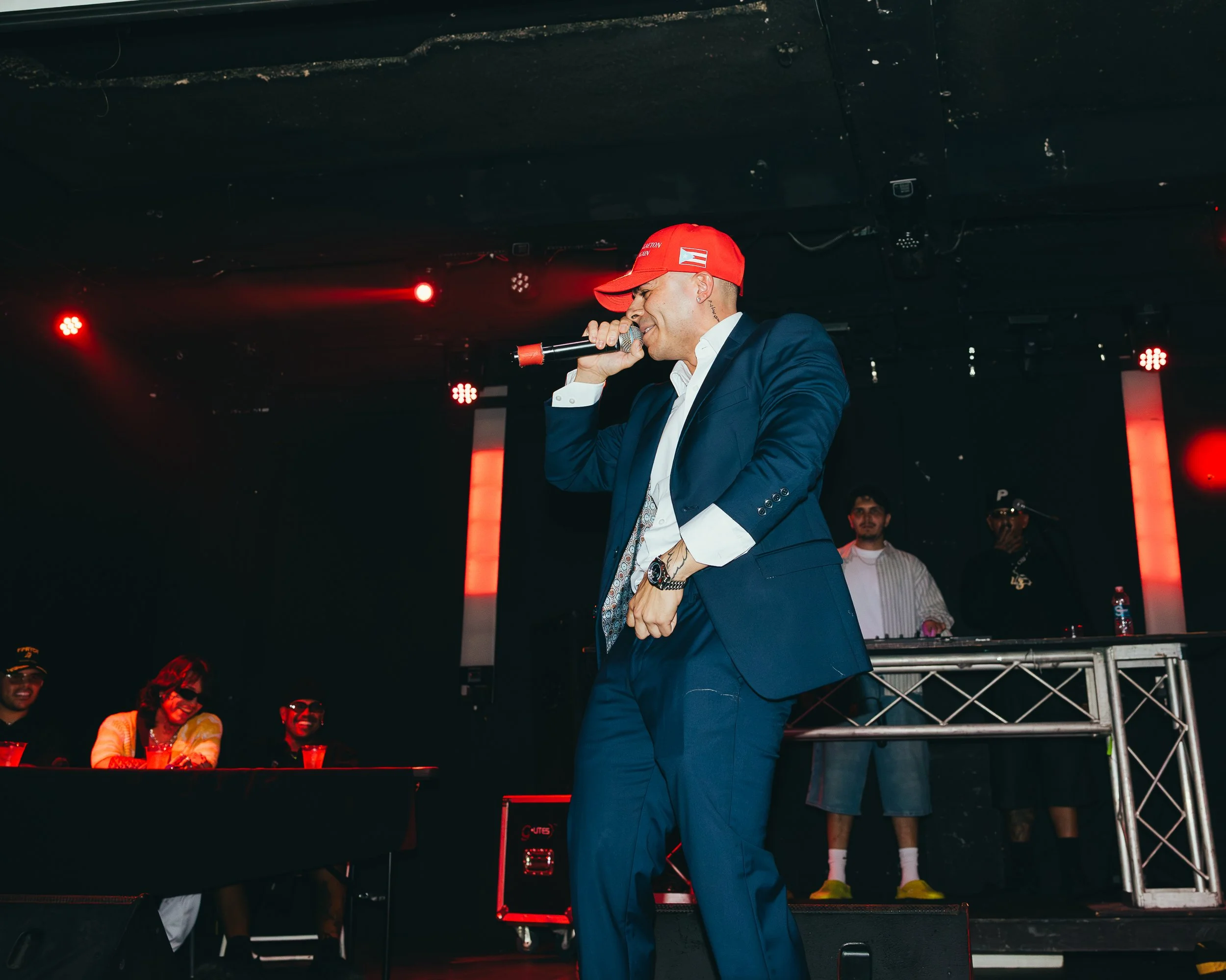Man wearing a blue suit and red cap performing with a microphone on stage, with a DJ and audience members visible in the background.