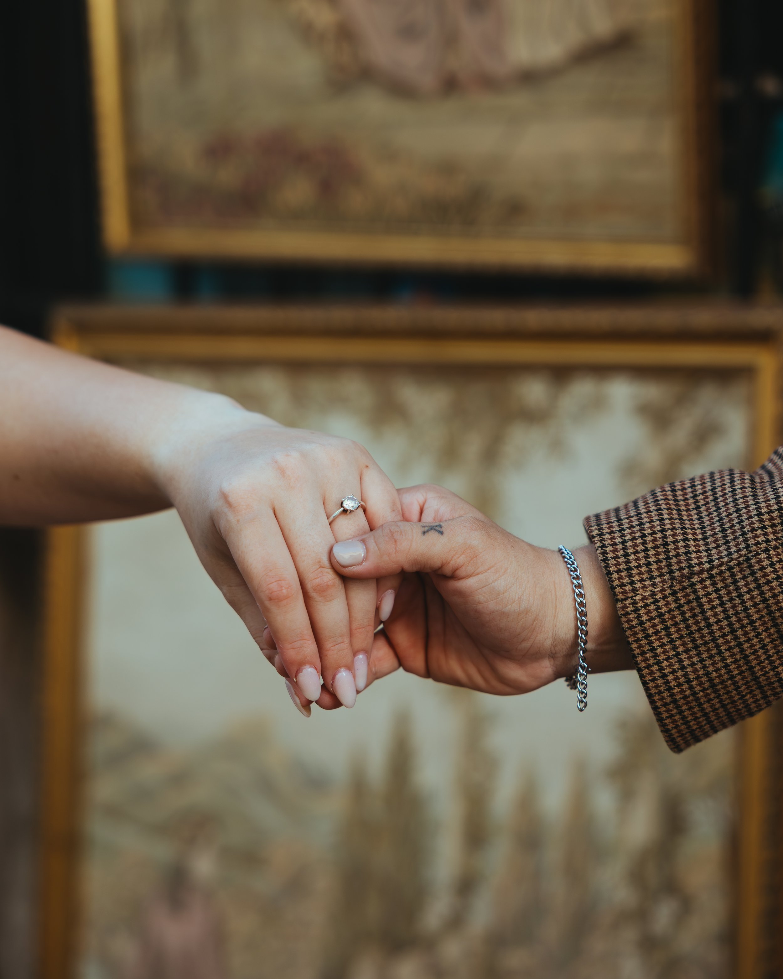 Close-up of a woman and man holding hands, with the woman showing her engagement ring; the woman has neatly manicured nails, and the man wears a chain bracelet and a brown checkered blazer, with a blurred background of paintings.