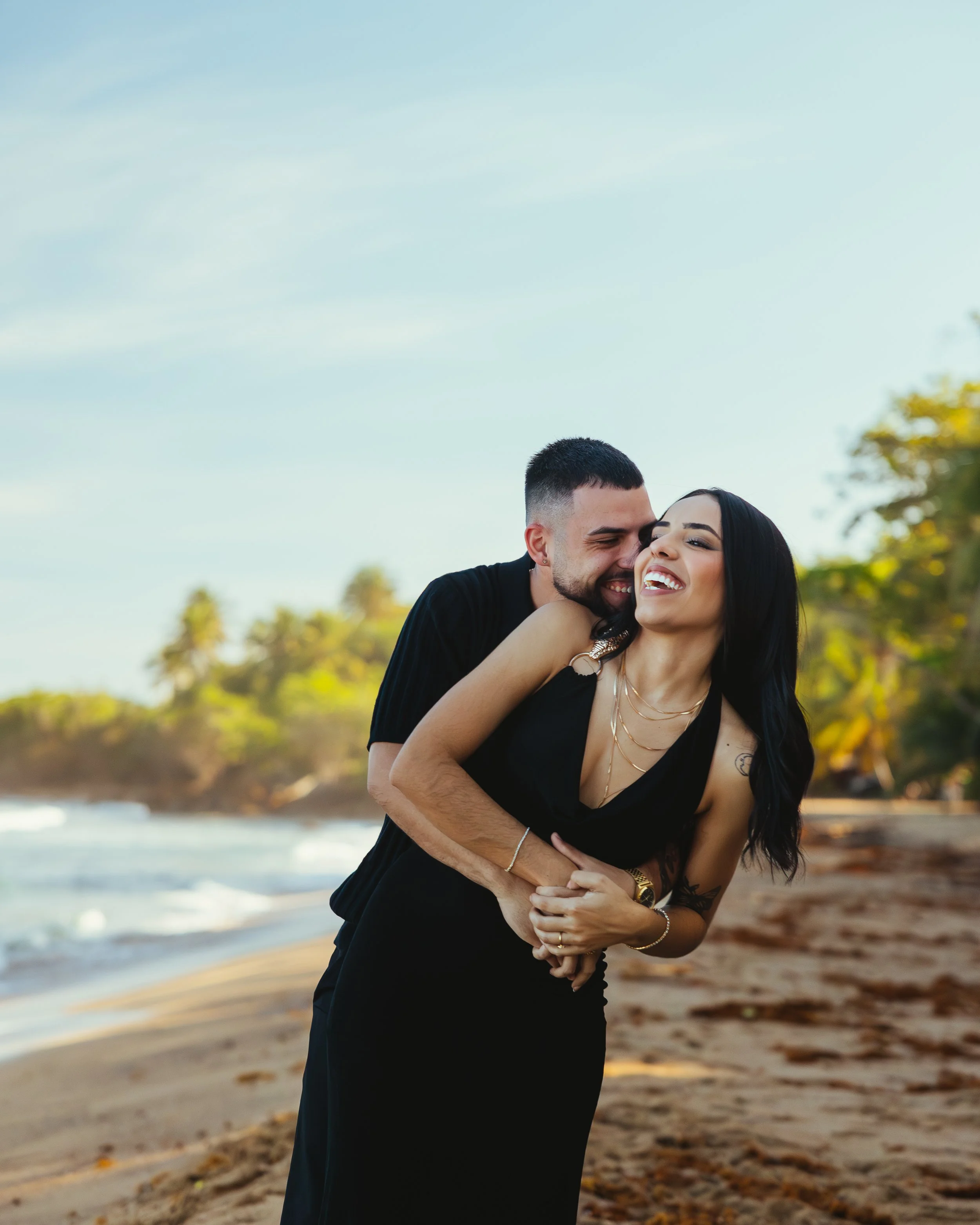 A couple enjoying a playful moment on a beach; the man is hugging and whispering to the woman, both laughing, with ocean waves and trees in the background.