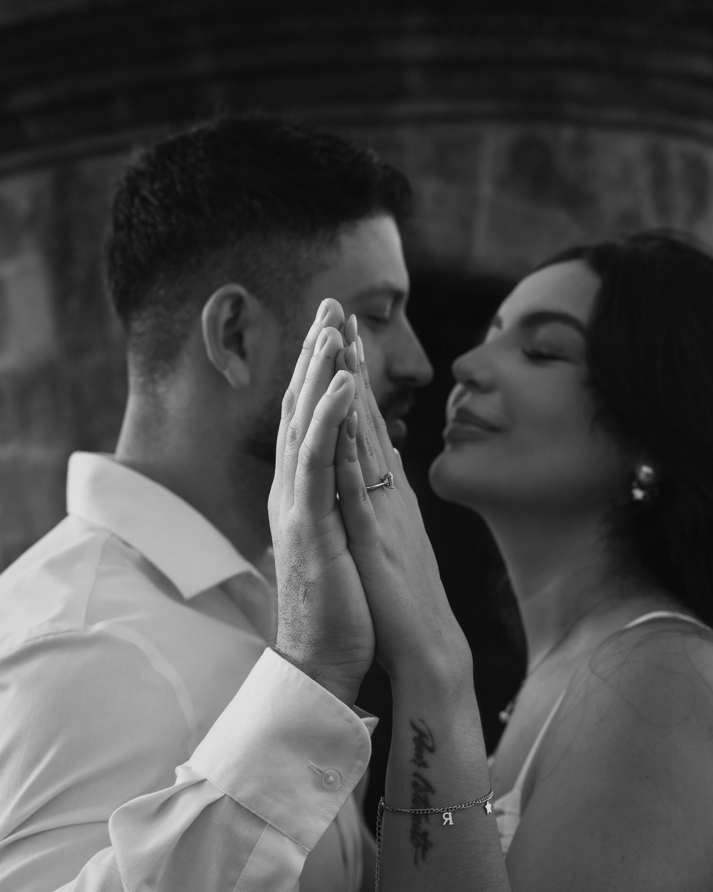 A black and white photo of a couple with their eyes closed, facing each other closely, holding hands with fingers intertwined, smiling gently.