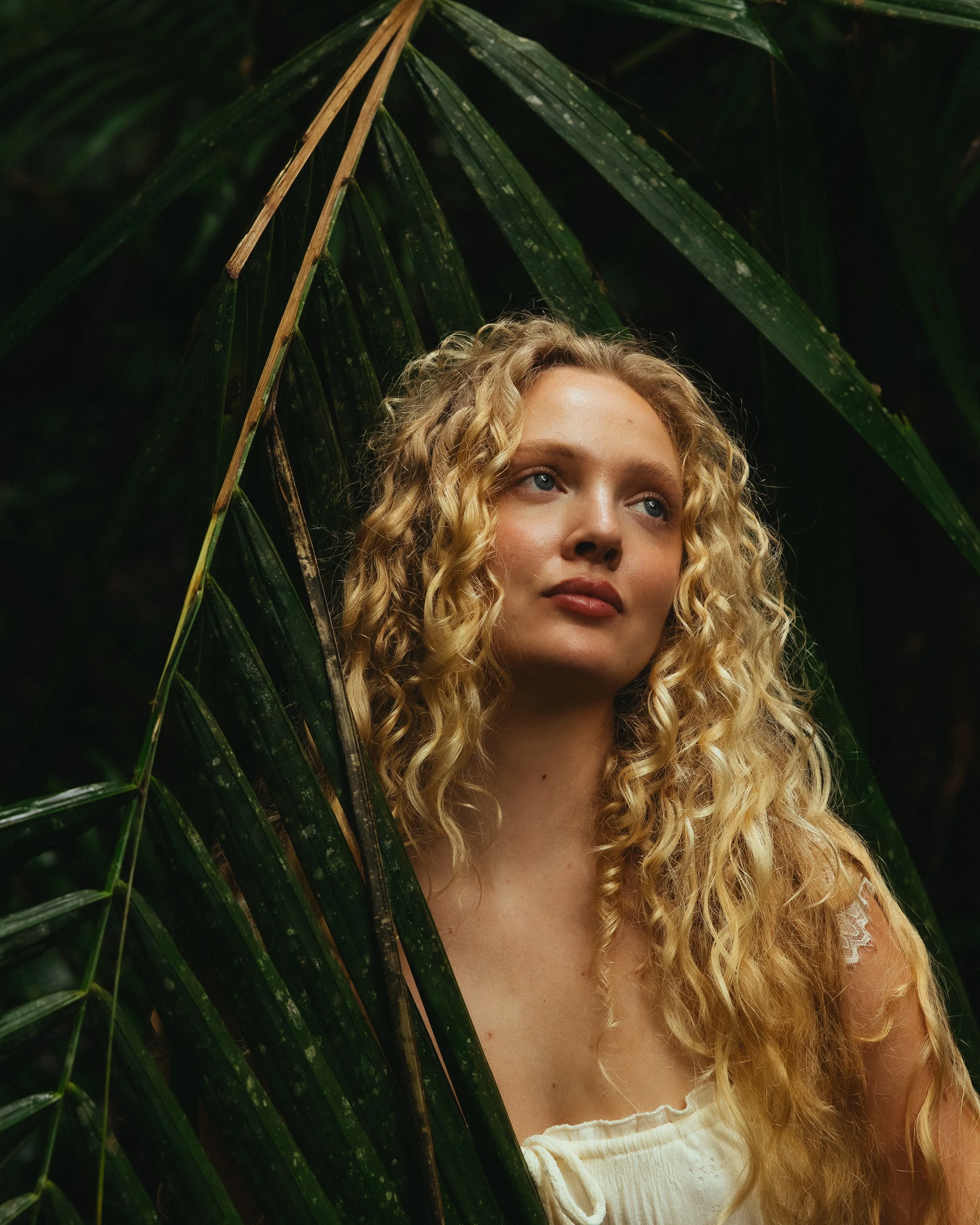 A woman with curly blonde hair and blue eyes peering from behind large green tropical leaves.