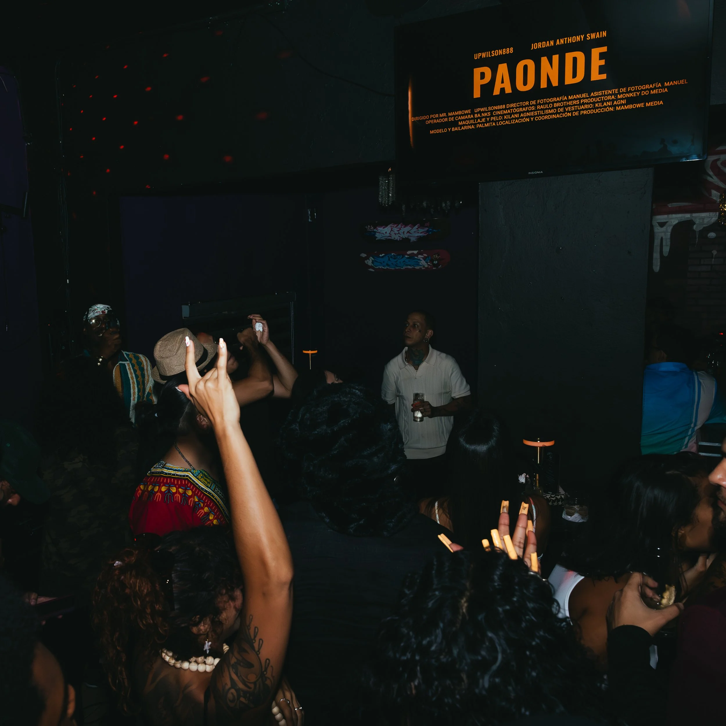 Crowd of people at a music event in a dark venue, with a man holding a drink on stage, and a large screen displaying the word 'PAONDE' along with artist and event details in bright orange text.