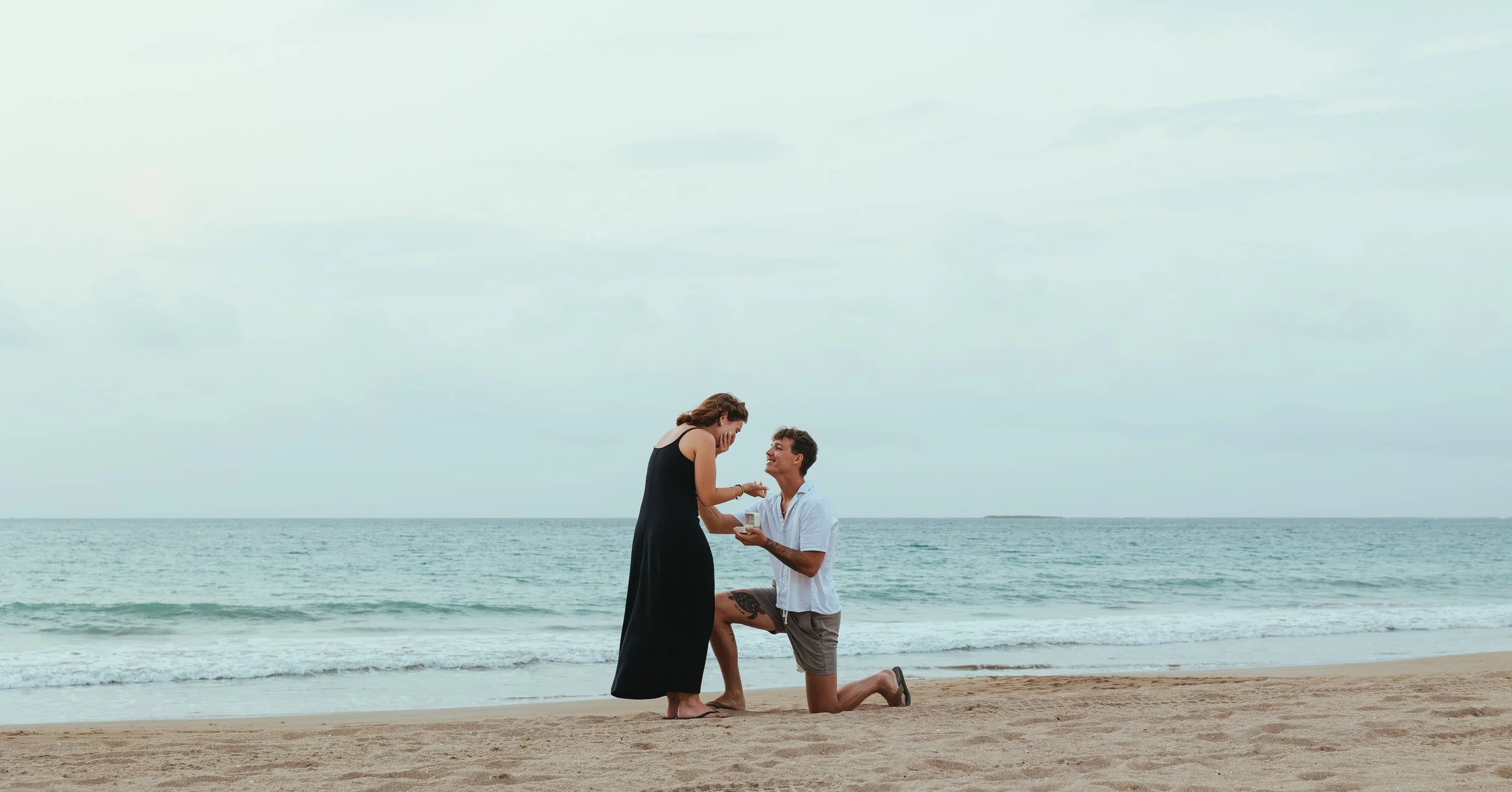 A man is proposing to a woman on a beach, with the man on one knee and holding a small box, as the woman leans forward with hands near her face, both smiling, with the ocean in the background.