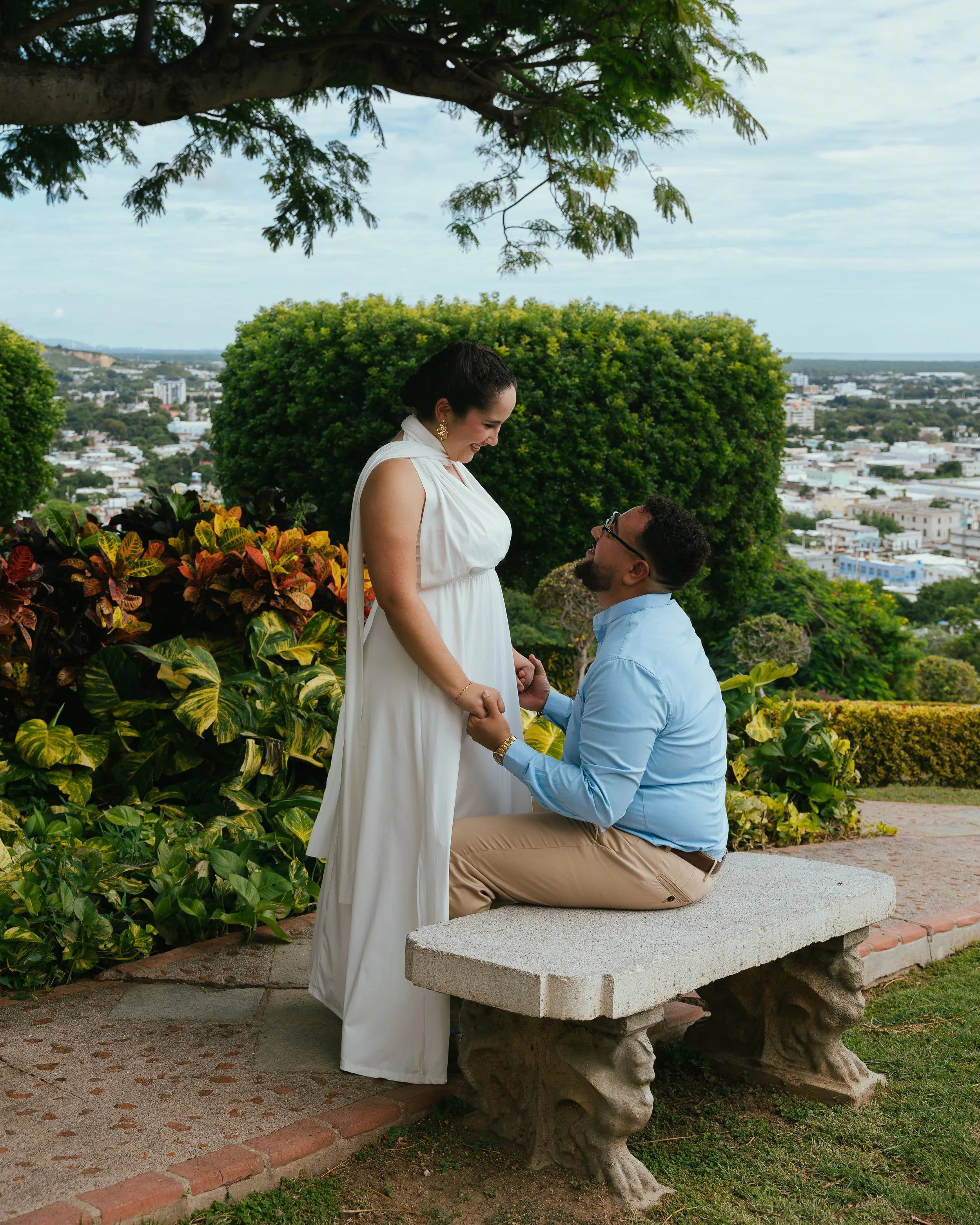 A man proposing to a pregnant woman on a park bench with city views in the background.