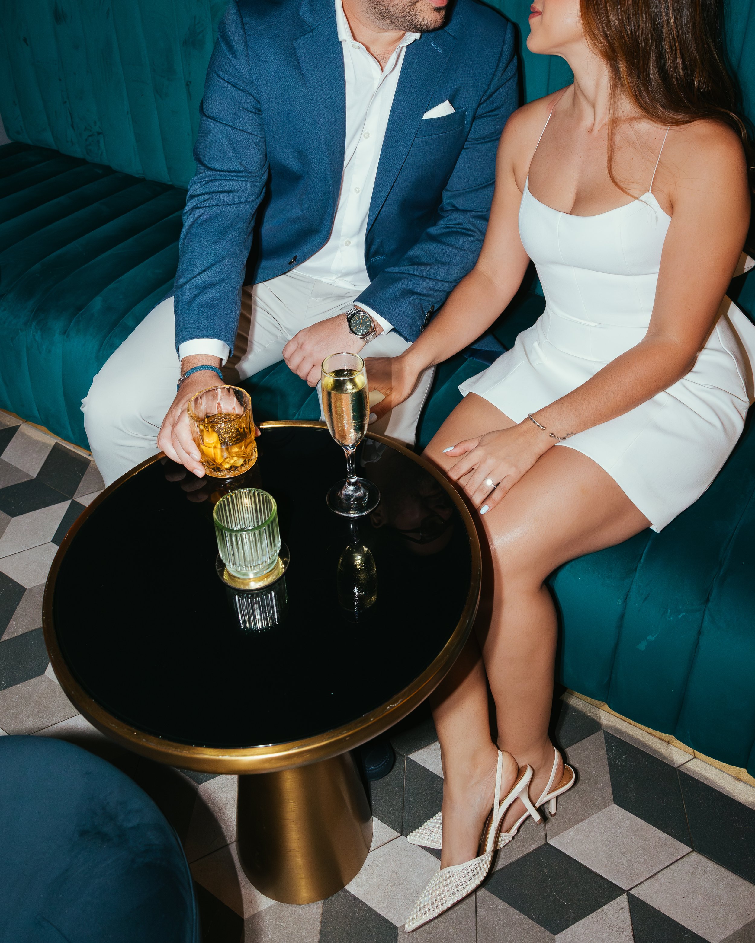 A man in a blue blazer and white pants, and a woman in a white dress, sitting on a teal velvet sofa, holding drinks on a black table, with the woman's legs crossed, wearing white high heels.