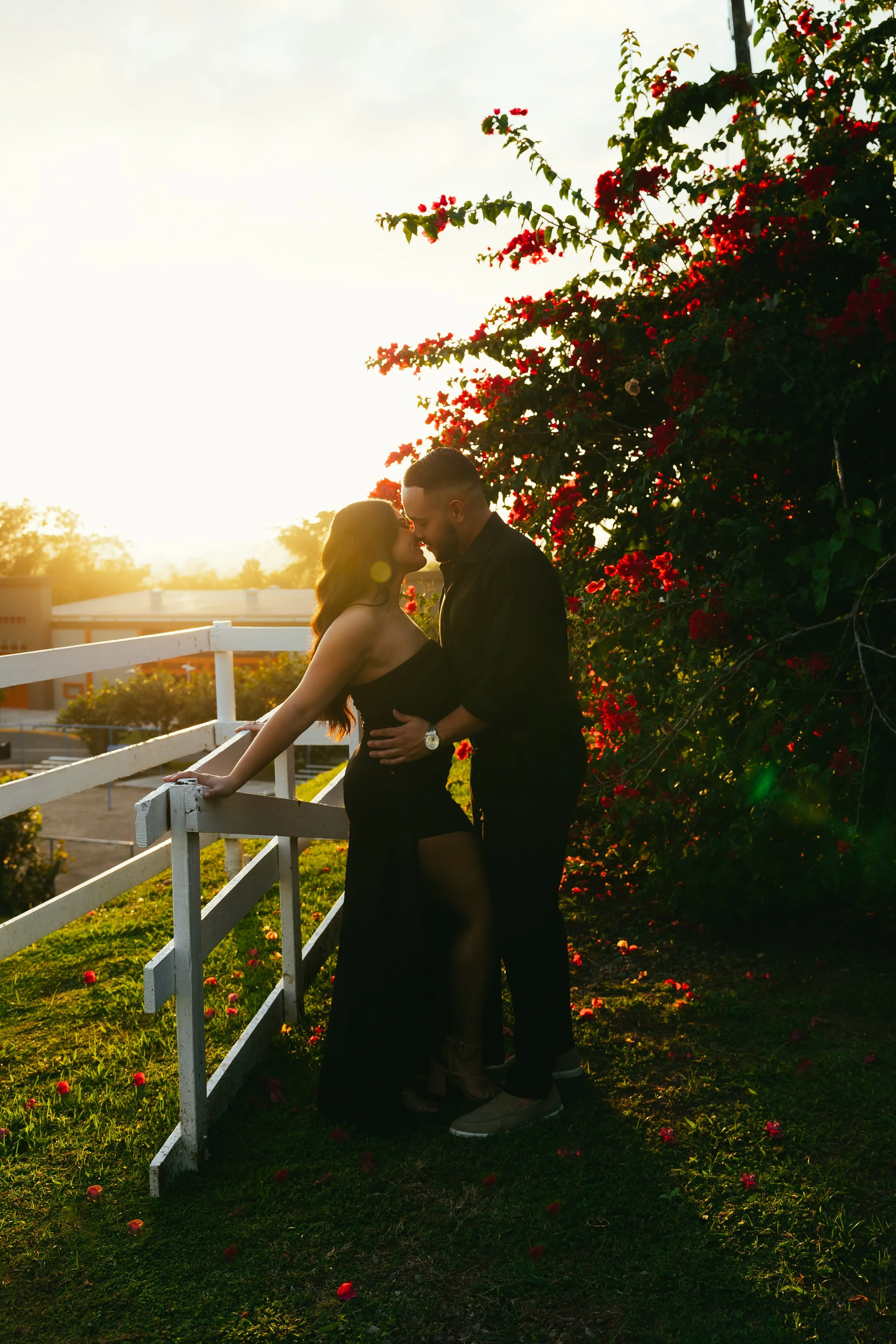 A romantic couple in love standing close together outdoors at sunset, with vibrant red flowers and greenery behind them, creating a warm and intimate scene.